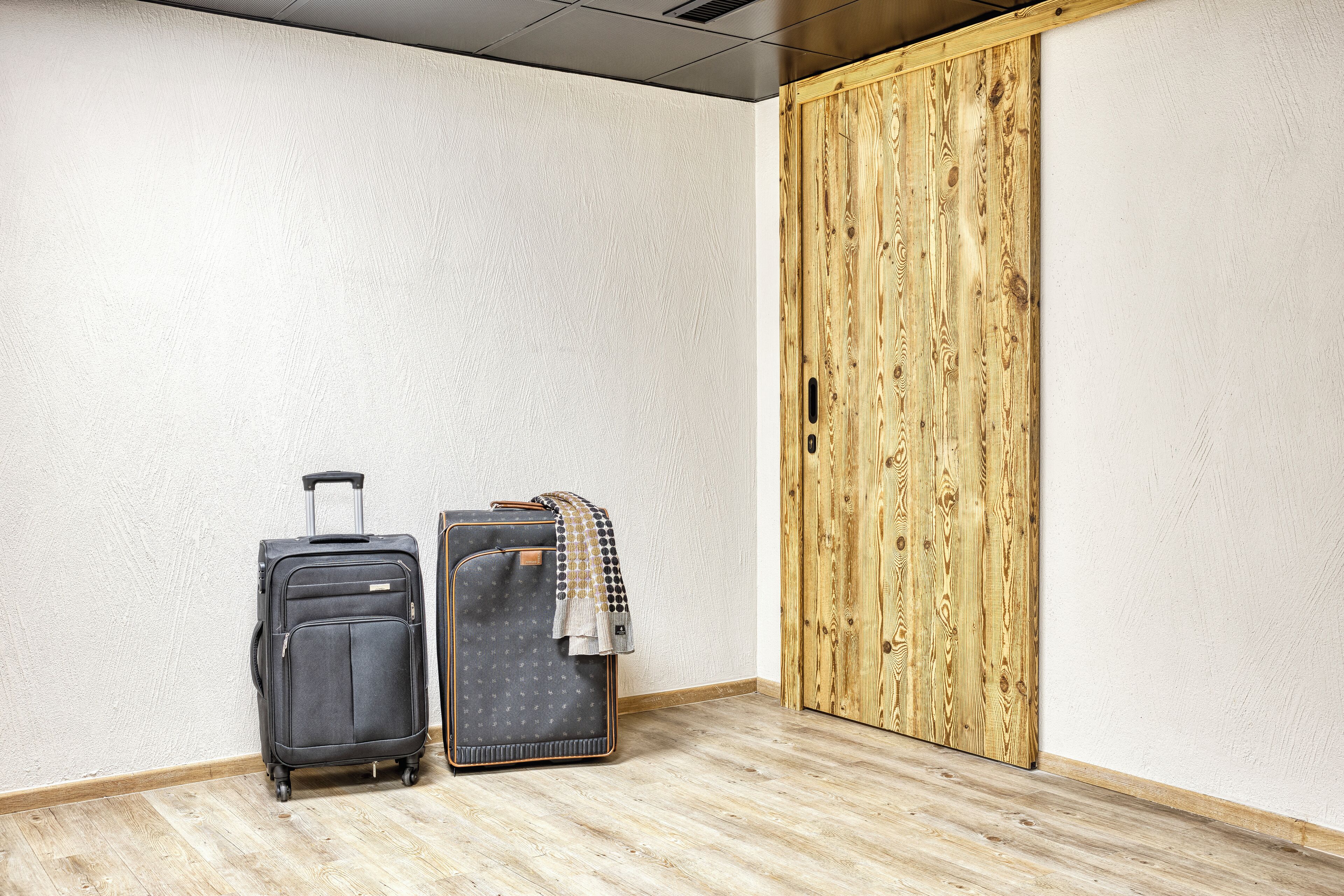 A room with a wooden door, a grey suitcase, and a patterned suitcase against a white wall.