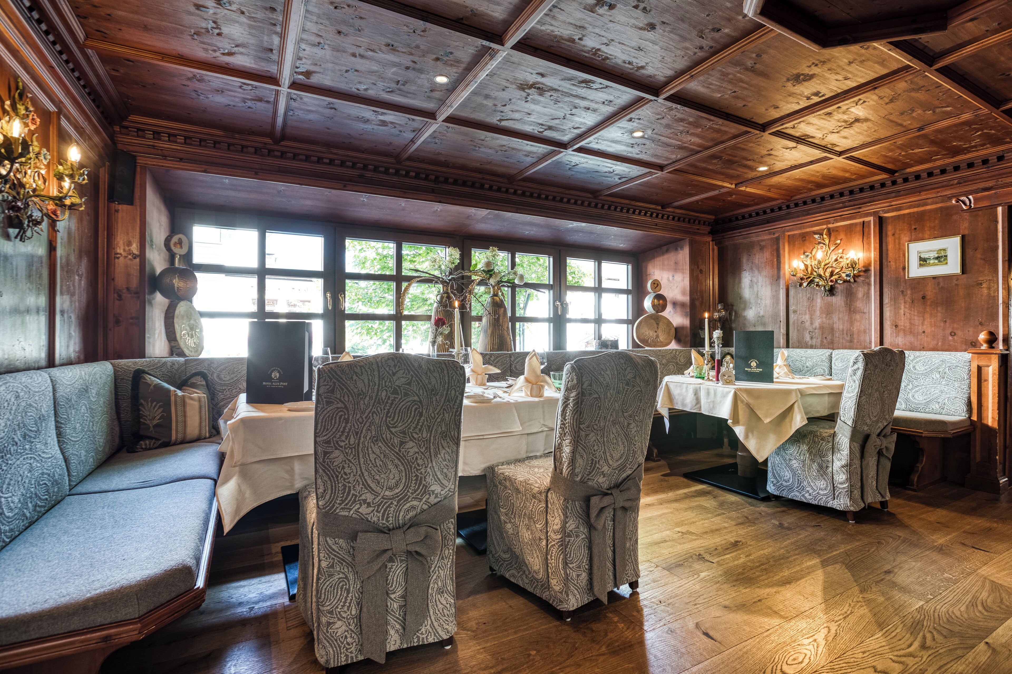 A dining area with a wooden ceiling, patterned chairs, a sofa, and a view through large windows.