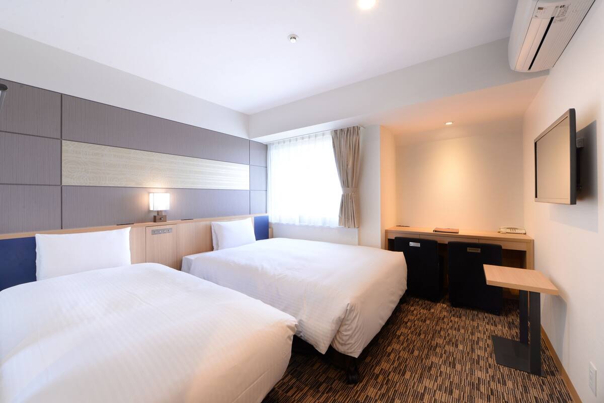 A hotel room with two beds, a desk, a chair, a TV, and a window with curtains.