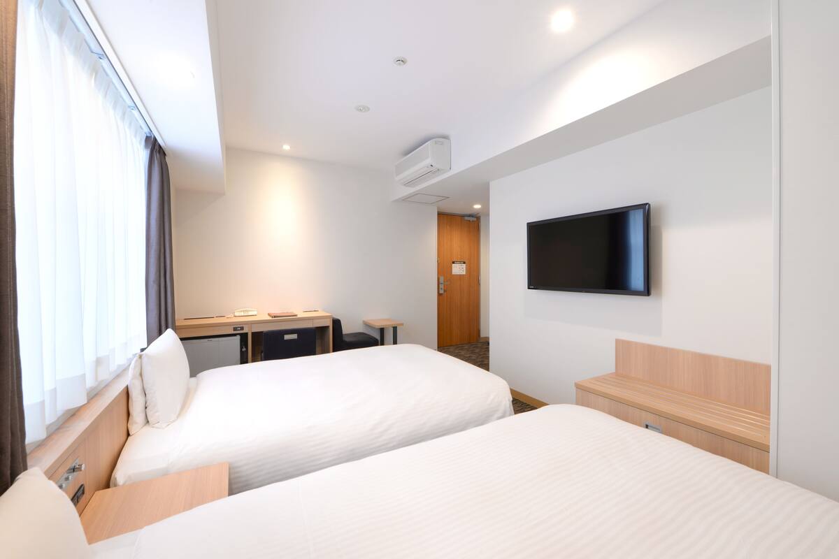 A hotel room with two beds, a desk, a chair, a television, and an air conditioning unit.