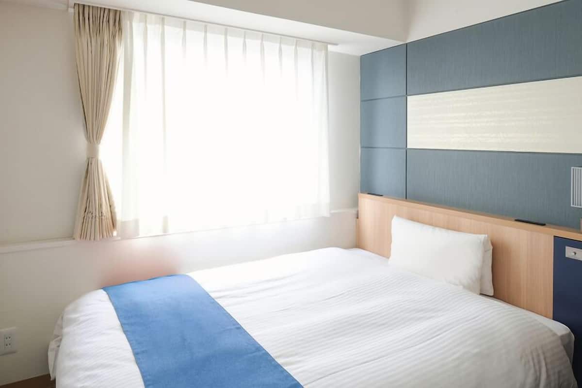A hotel room with a bed, a TV mounted on the wall, a bedside table with a lamp, and a window with curtains.