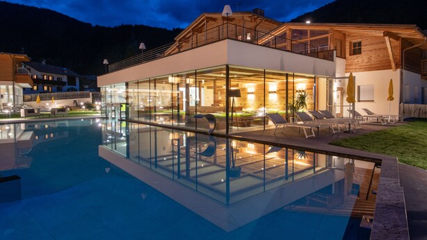 Un hotel moderno con piscina de vidrio, mobiliario exterior y fachada de madera.