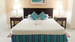 Una cama bien hecha con almohadillas a rayas y una toalla doblada, dos lámparas de noche y un cuadro con una playa en la pared.