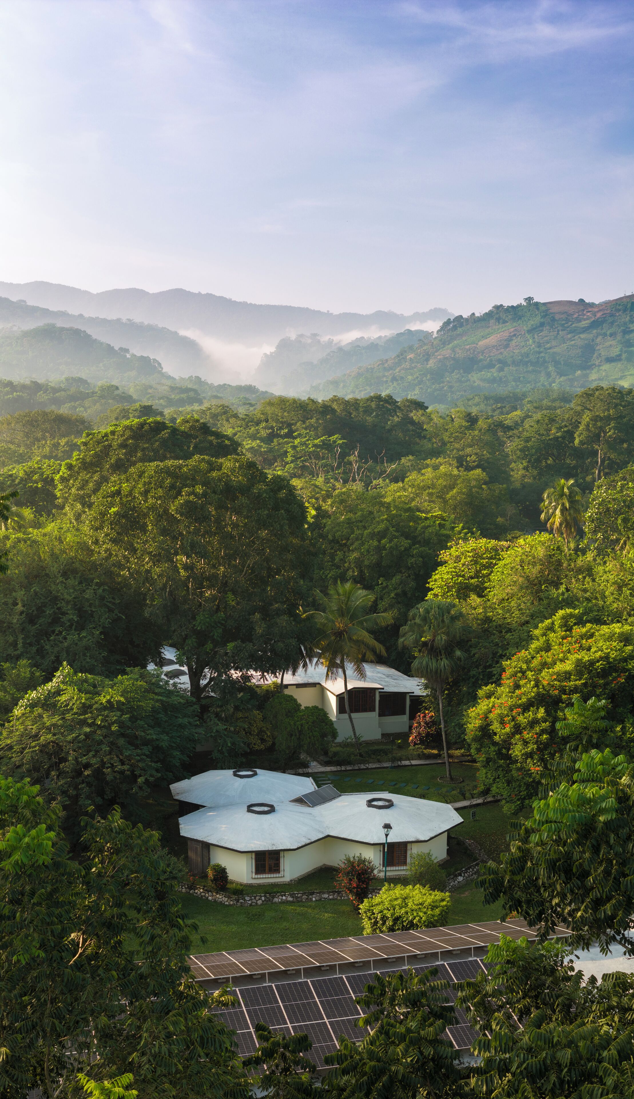 A house with a sloped roof and solar panels, surrounded by dense greenery and hills in the background.