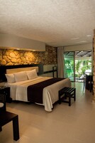 Un dormitorio con pared de piedra, una cama con ropa de cama blanca, una mesita de noche, un banco de madera y vistas a un jardín por una puerta de cristal.