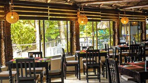 Un restaurante con techo de paja, mesas y sillas de madera, y amplios ventanales que dan a vistas de vegetación.
