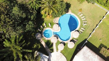 Vista aérea de una piscina rodeada de sillones de descanso y sombrillas, con exuberante follaje y una palmera al fondo.