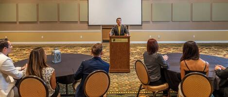 A conference room with a speaker at a podium, audience seated at round tables, and a large screen on the wall.