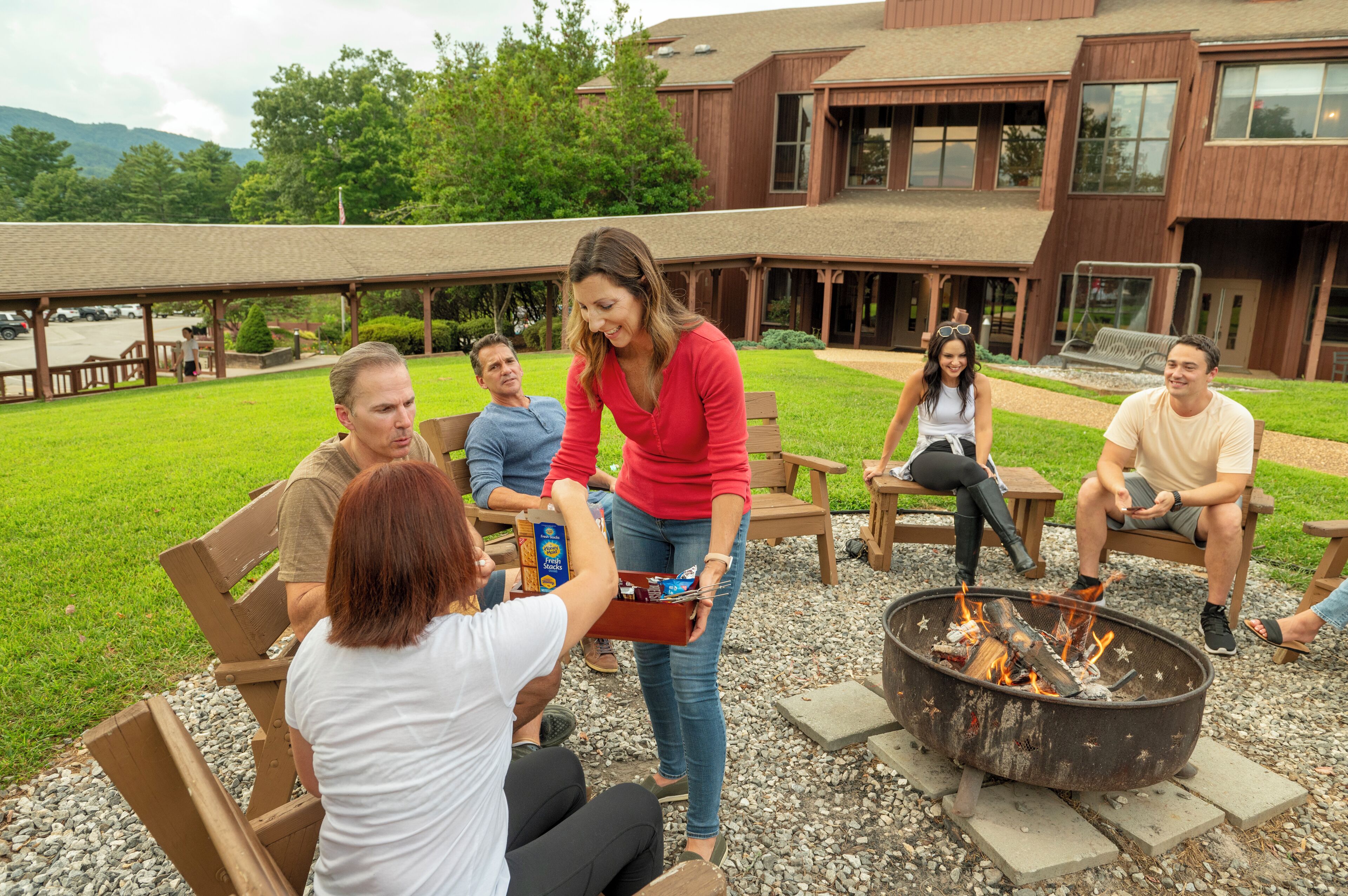A group of people sitting on wooden benches outdoors with a fire pit in the center.