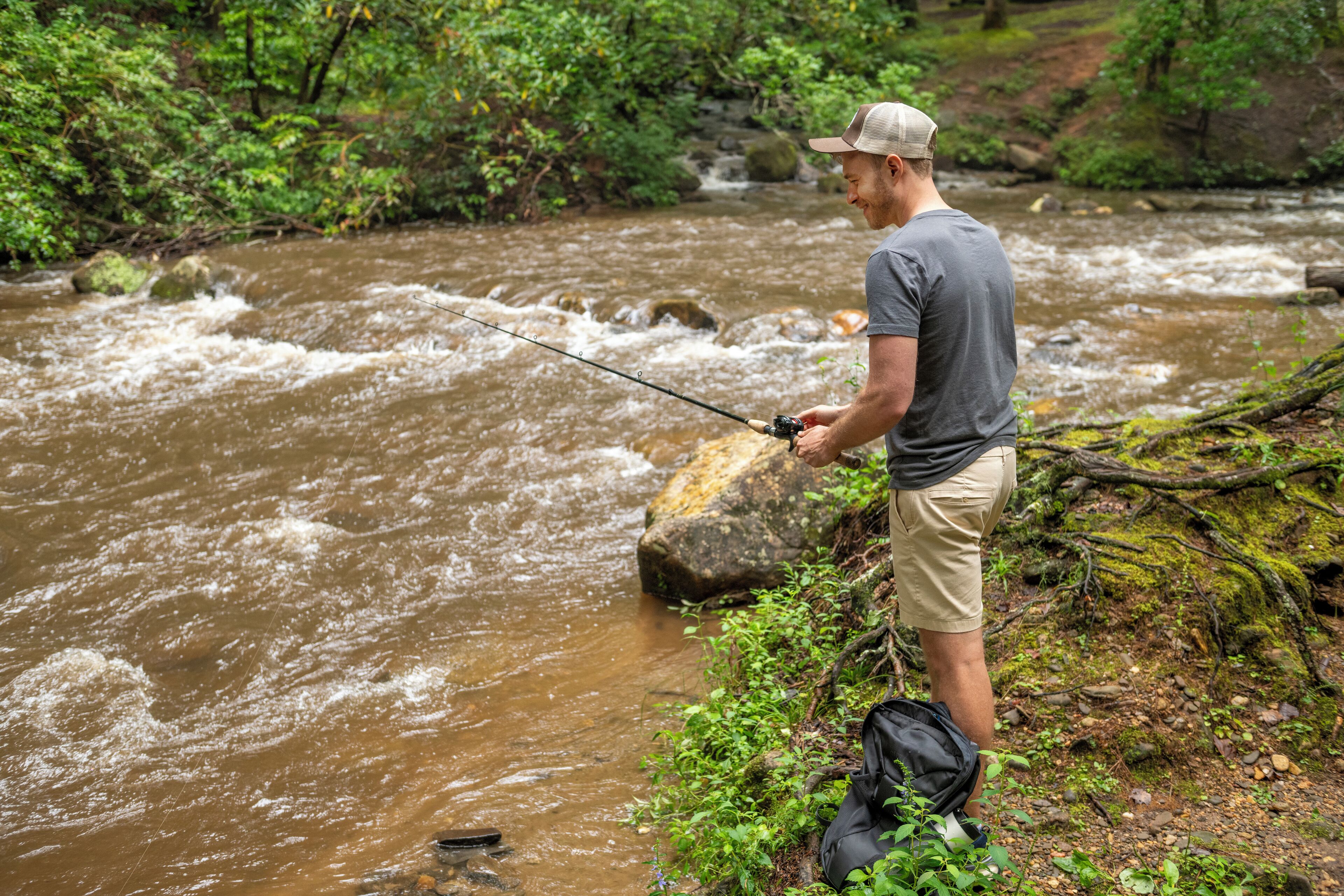 A man fishing by a river with a backpack on the ground.