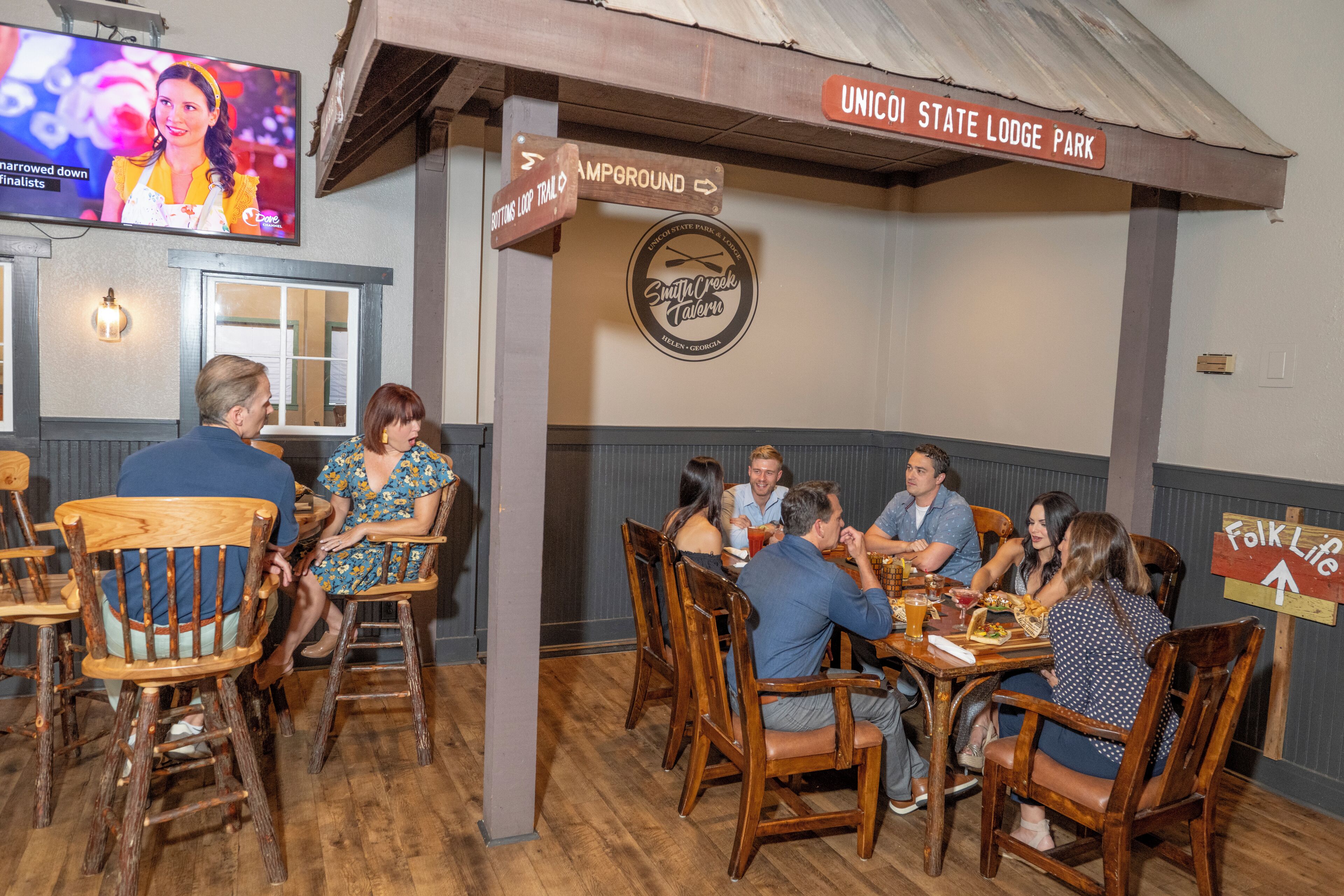 Un restaurante con sillas y mesas de madera, un televisor montado en la pared y señales que indican el camino a atracciones cercanas.