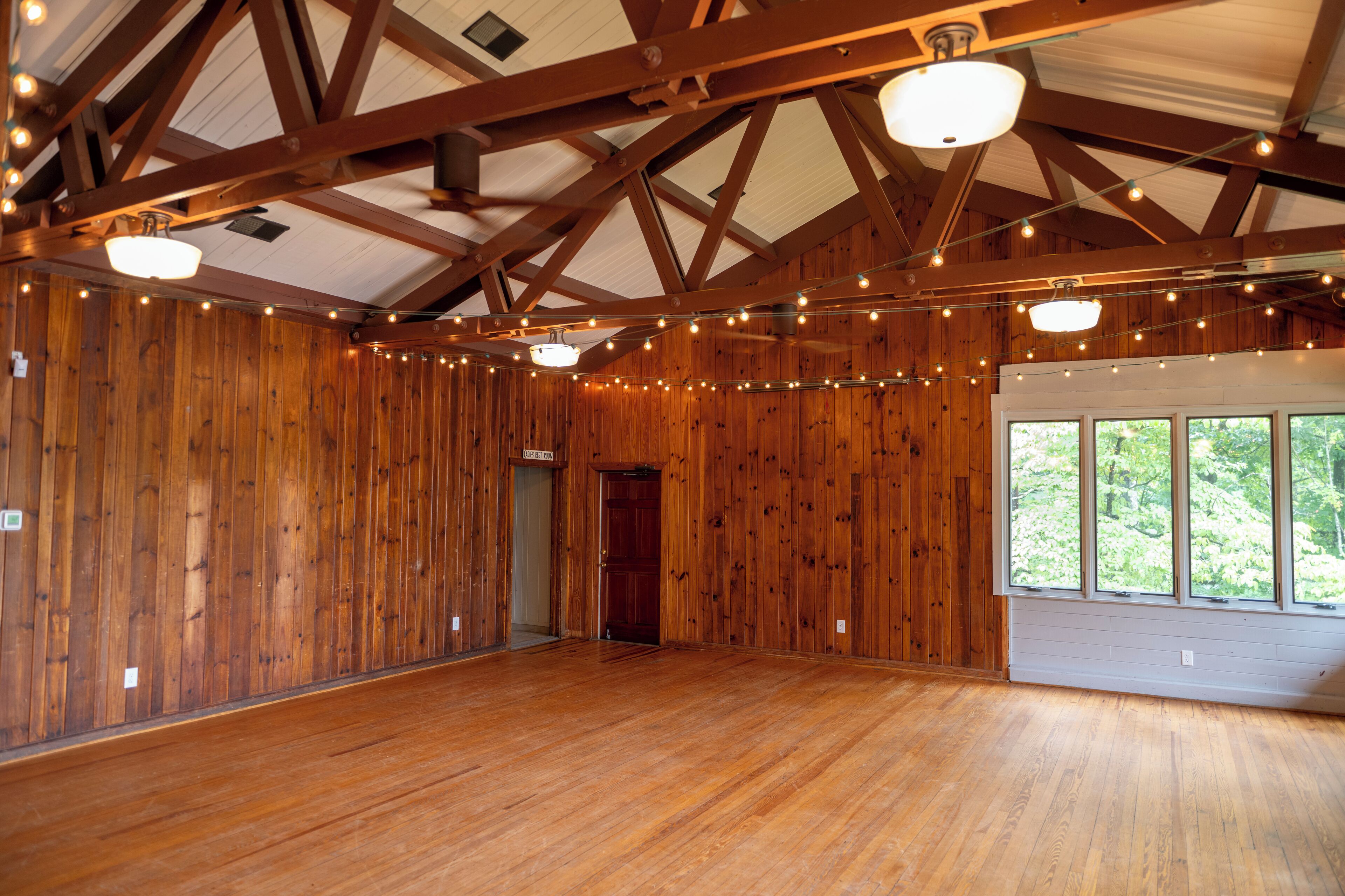 A spacious room with wooden walls and a high ceiling supported by wooden beams. There are string lights hanging from the ceiling, and a door leading to another area.