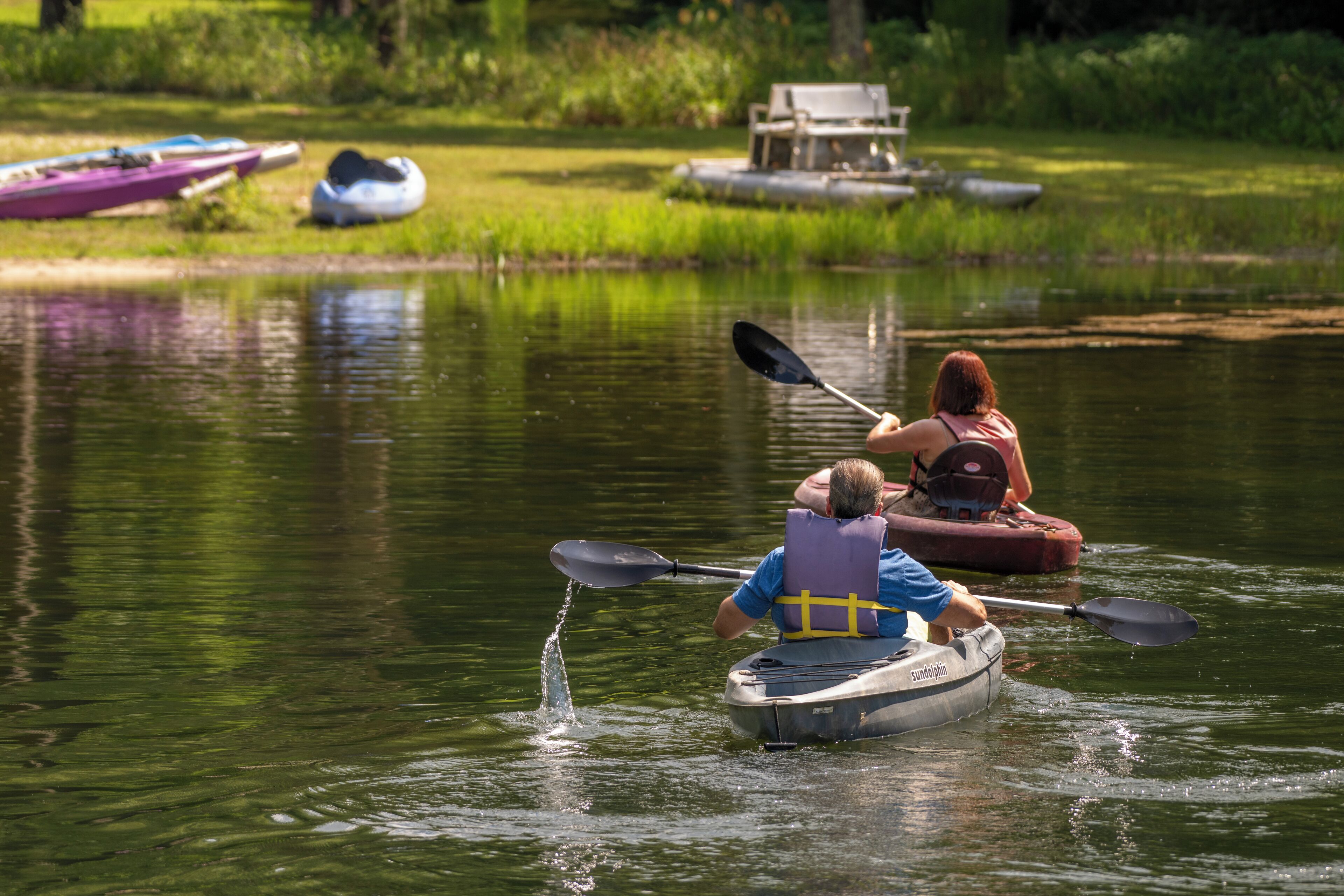 A person kayaking on a calm river with another person in a rowboat nearby.