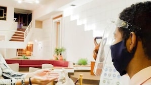 Una persona con protector facial entrega una botella a otra persona sentada en una mesa, en un espacio interior bien iluminado con una escalera y un sofá rojo al fondo.