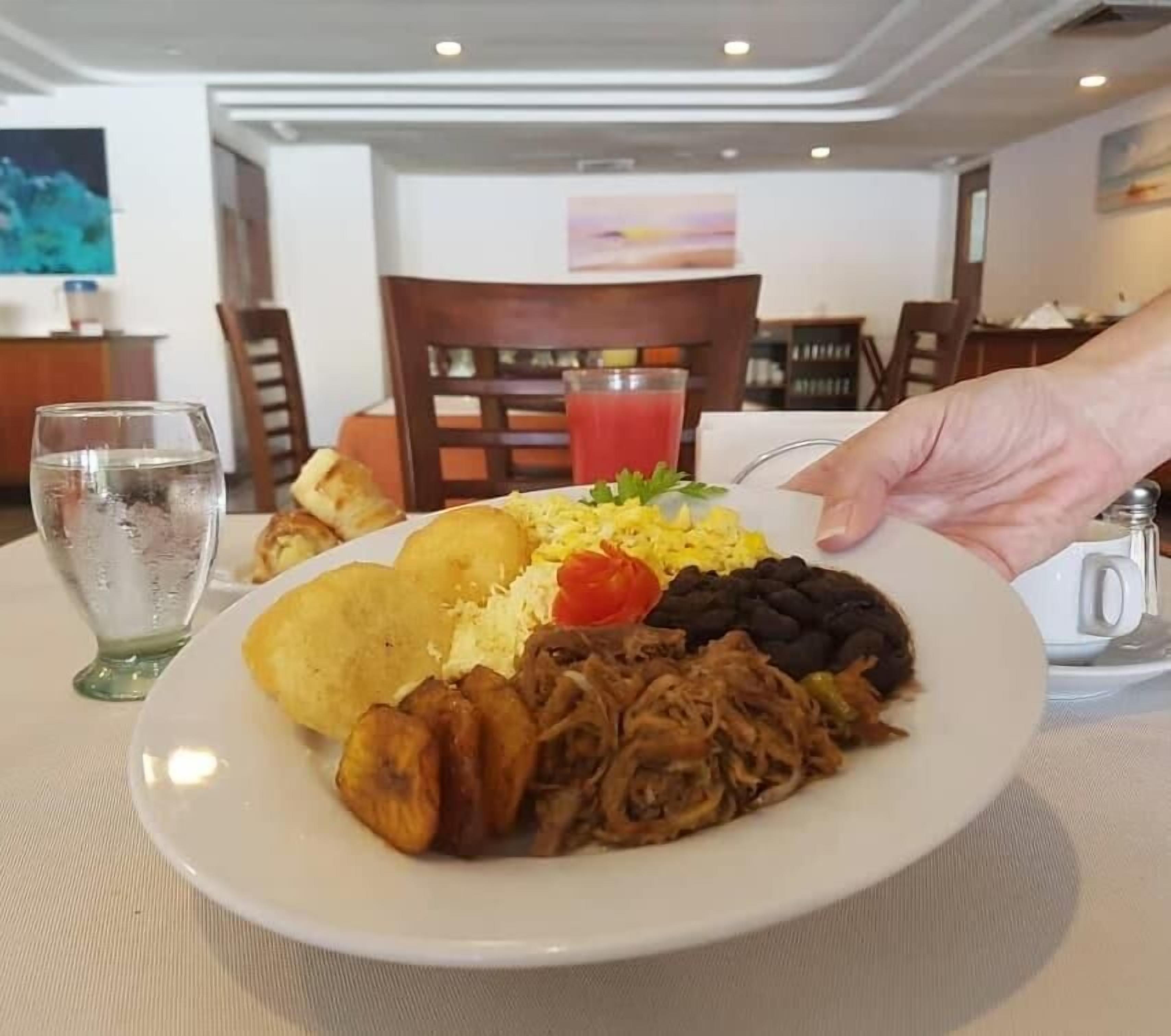 Un plato de comida con arroz, frijoles, carne, un vaso de agua y una rebanada de pan sobre una mesa en un comedor.