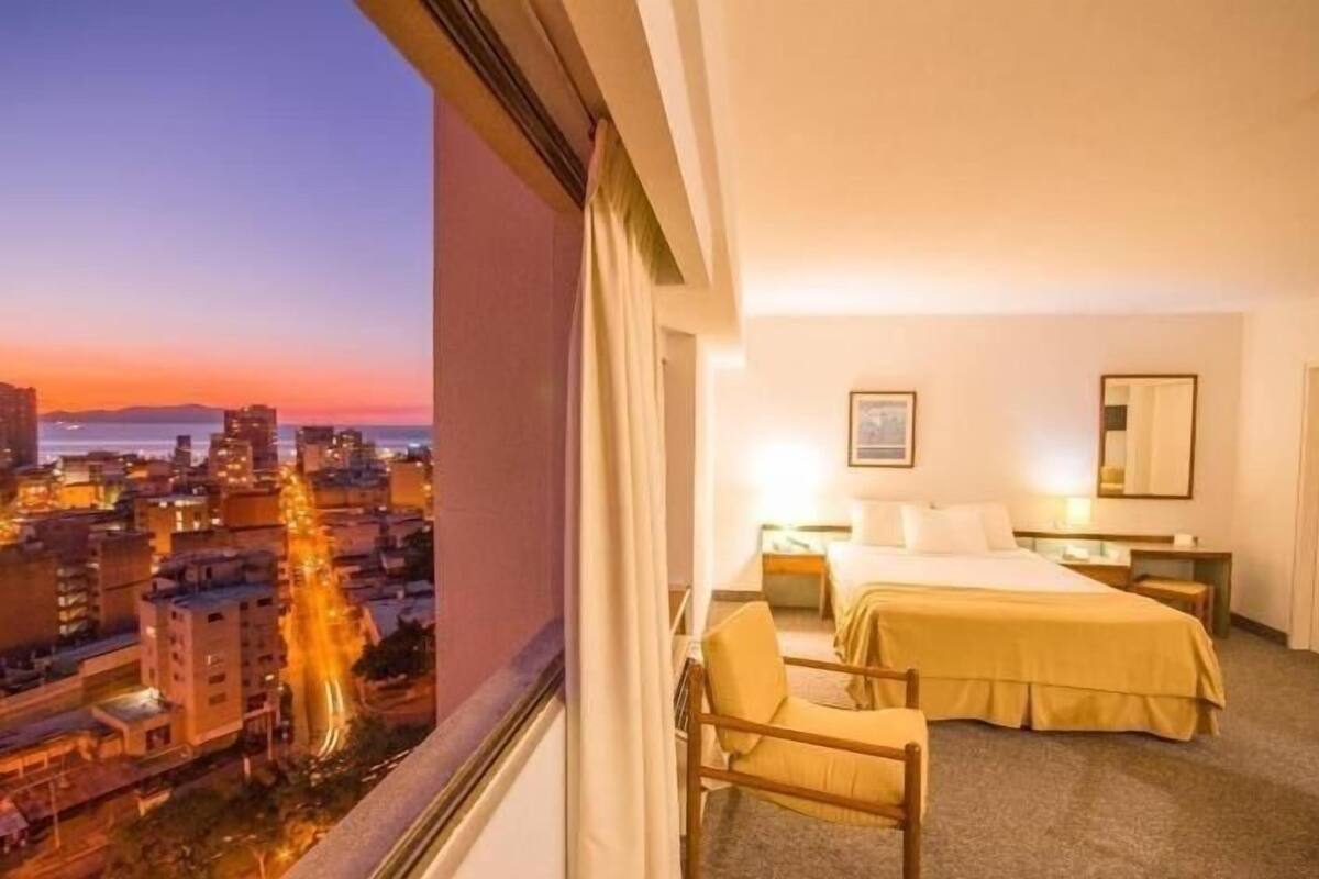 A hotel room with a bed, bedside table, chair, and a city view.