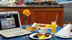 Una mesa con desayuno: un ordenador portátil, varias frutas, bollería y una bebida.