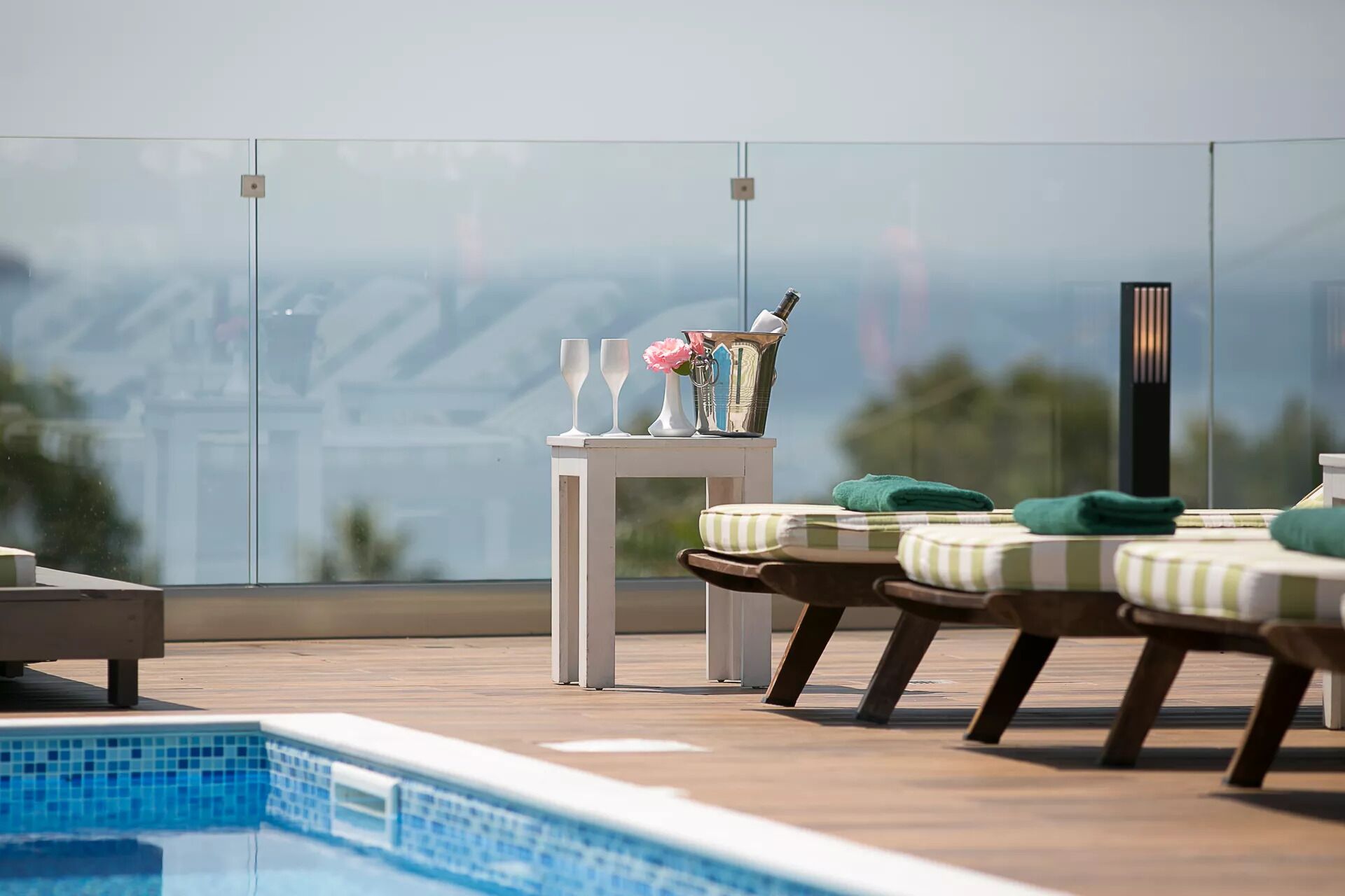 A rooftop pool area with lounge chairs, a small table with a bottle and flowers, and a view of the mountains.