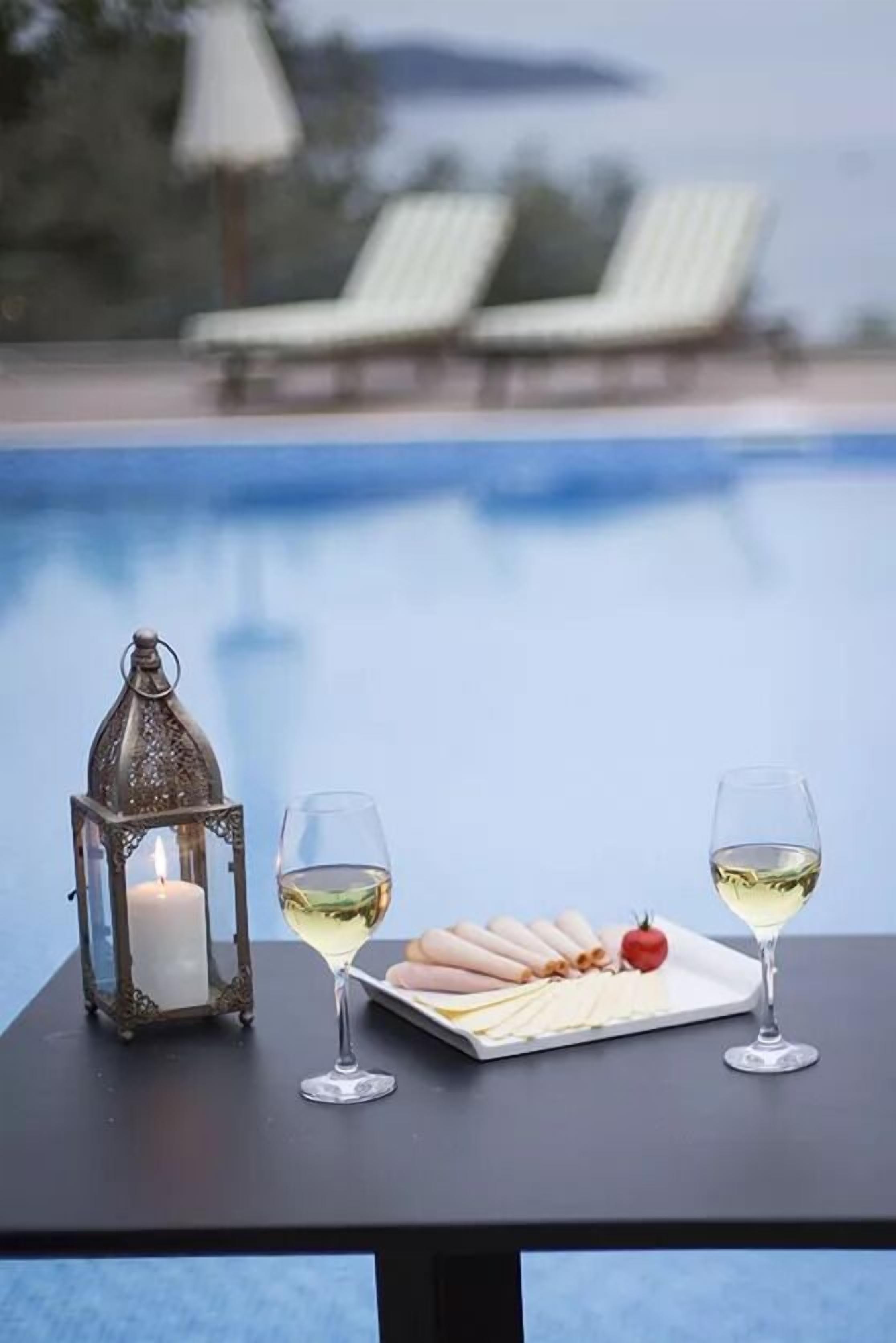 A table with a candle, two glasses of white wine, and a plate of food by a poolside.