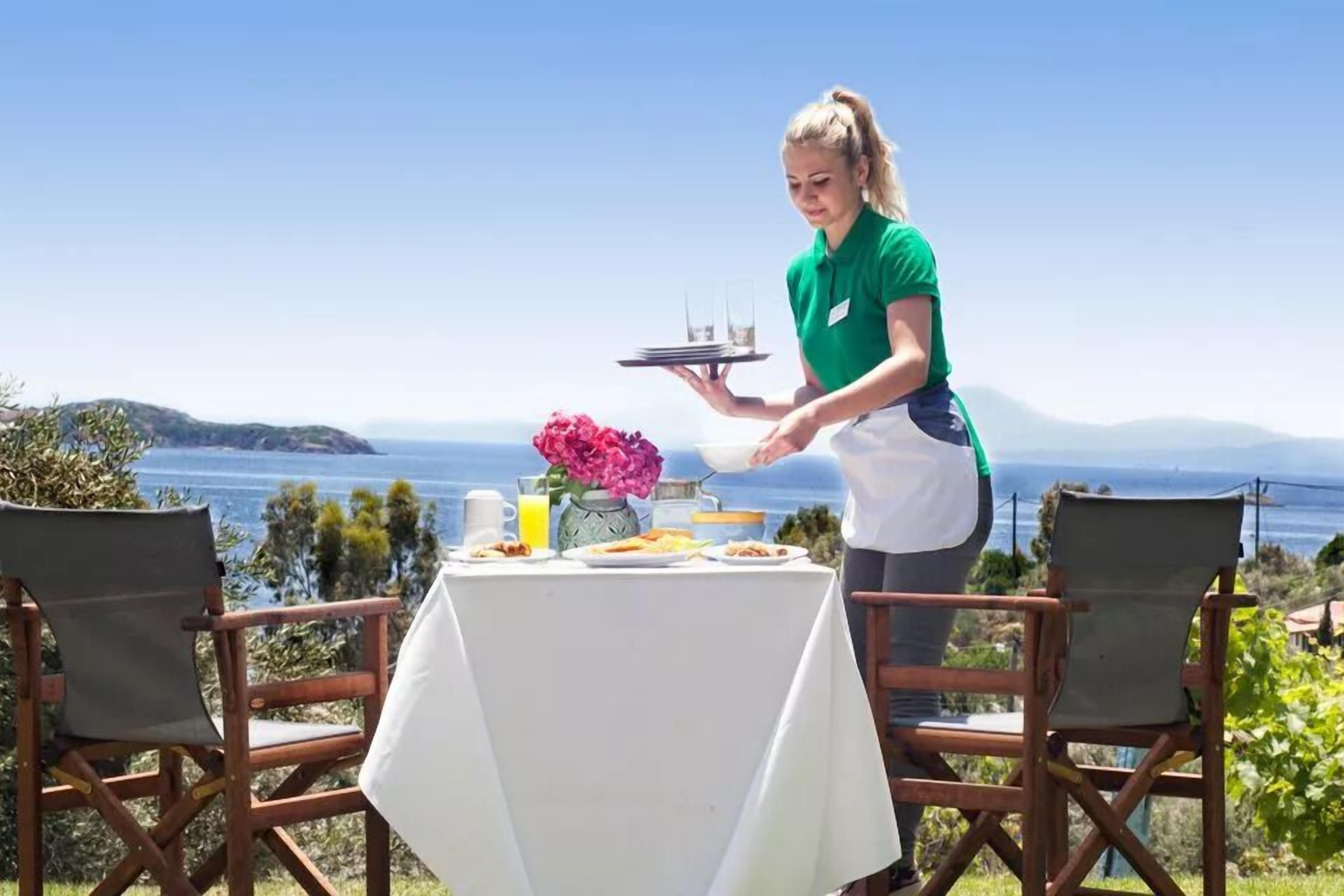 Tavola imbandita per la colazione con vista sul mare, un cameriere al servizio e un vaso di fiori.