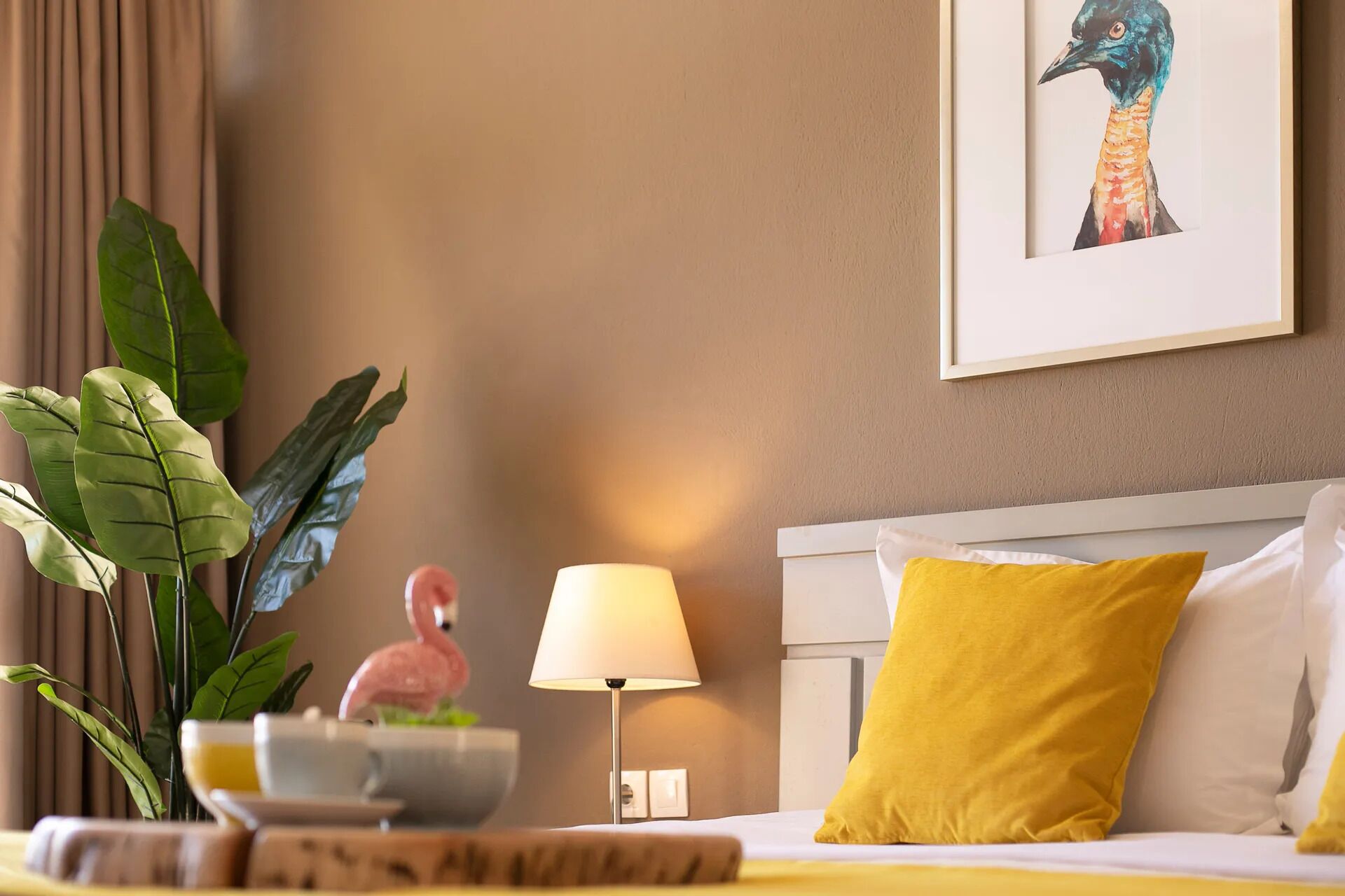 A hotel room with a bed, a nightstand, a lamp, a plant, and a framed picture on the wall.