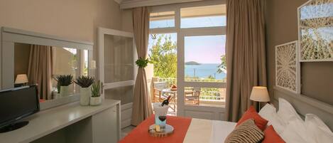 Quarto de hotel com cama, escrivaninha com televisão, varanda com mesa e cadeiras e vista para o mar.