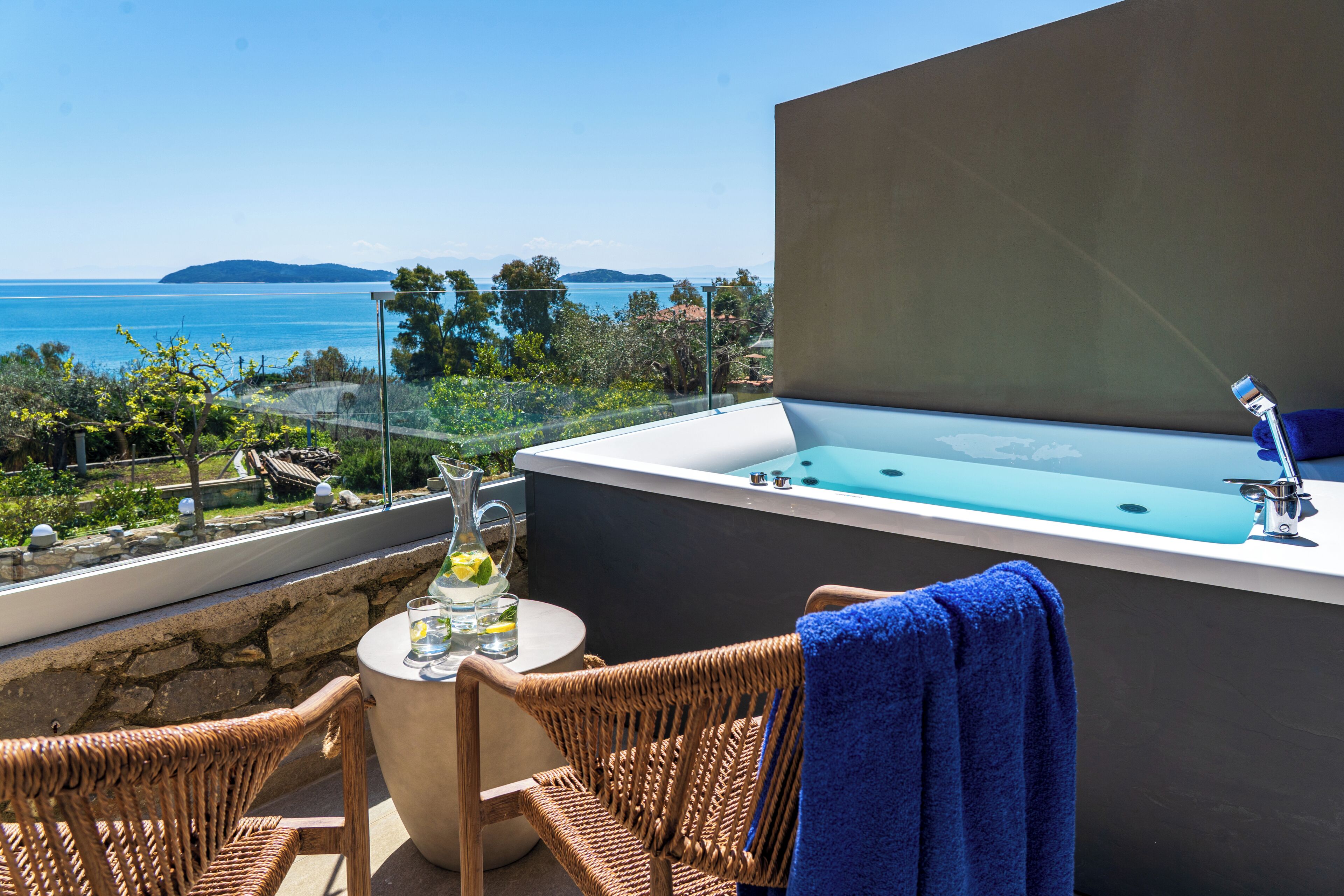 Un balcón con vista al mar, una bañera, muebles de mimbre y una mesa con un vaso de agua.