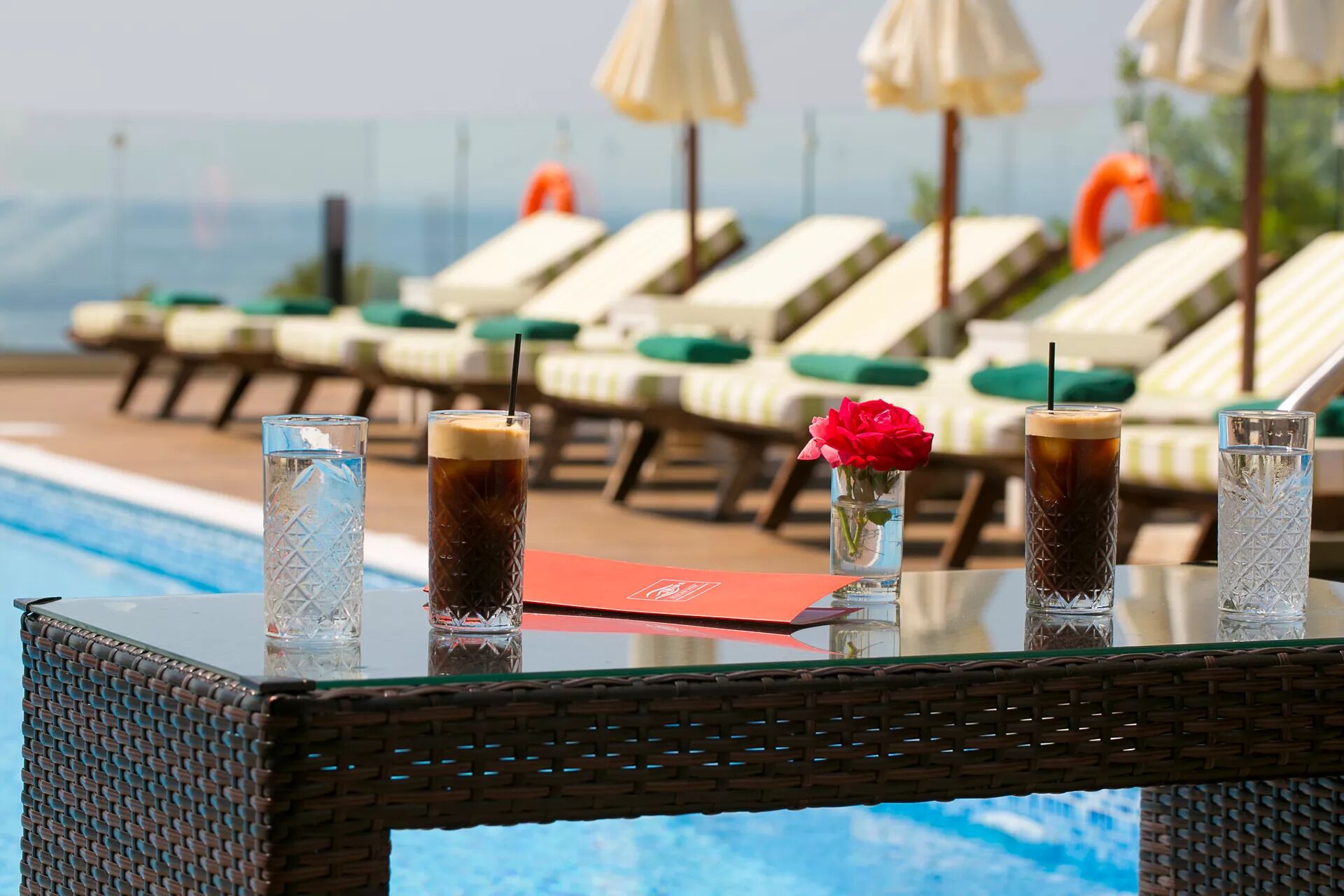 A poolside table with drinks and a red menu, surrounded by lounge chairs and umbrellas.