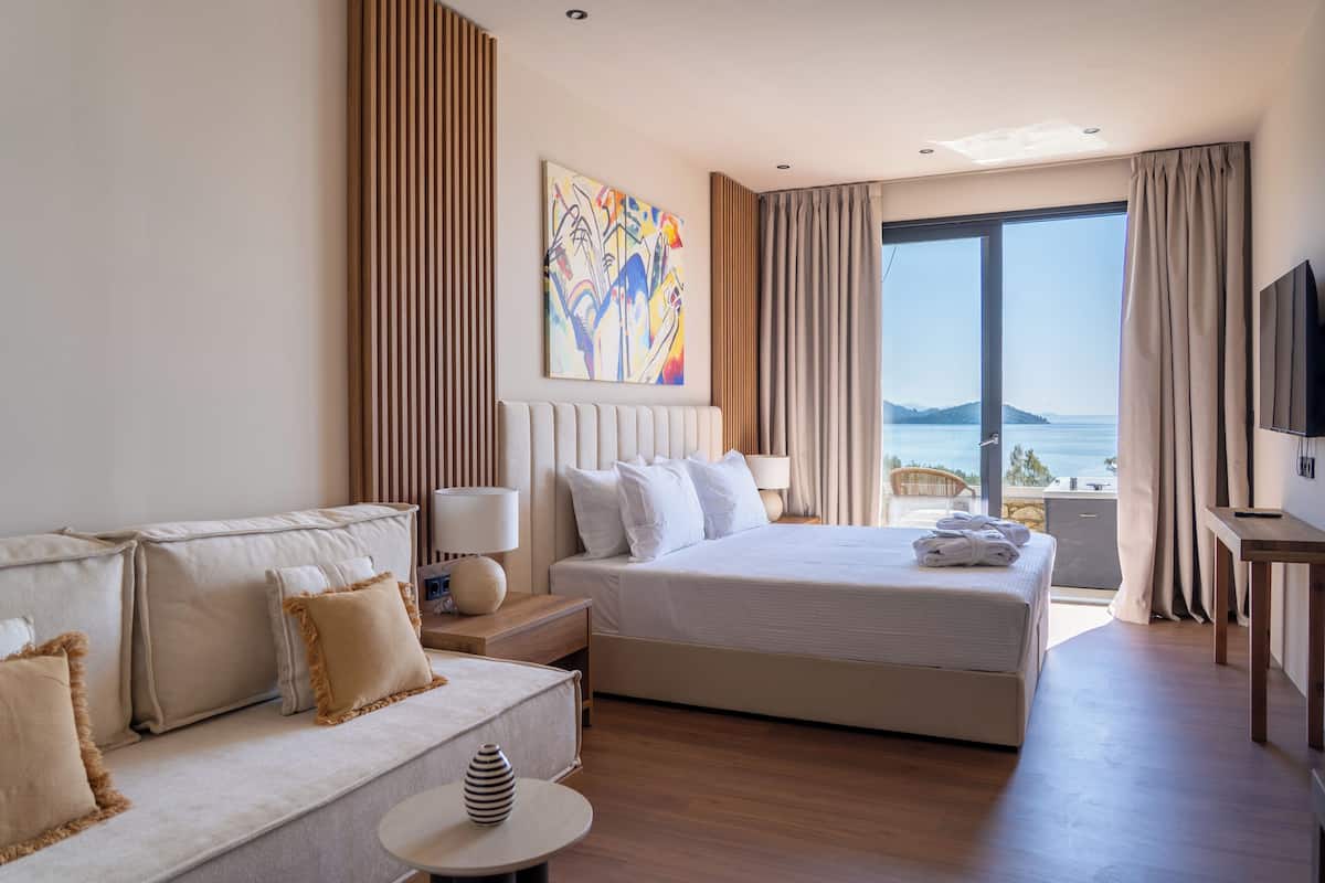 Habitación de hotel con cama, sofá, televisor y un amplio ventanal con vista al mar.
