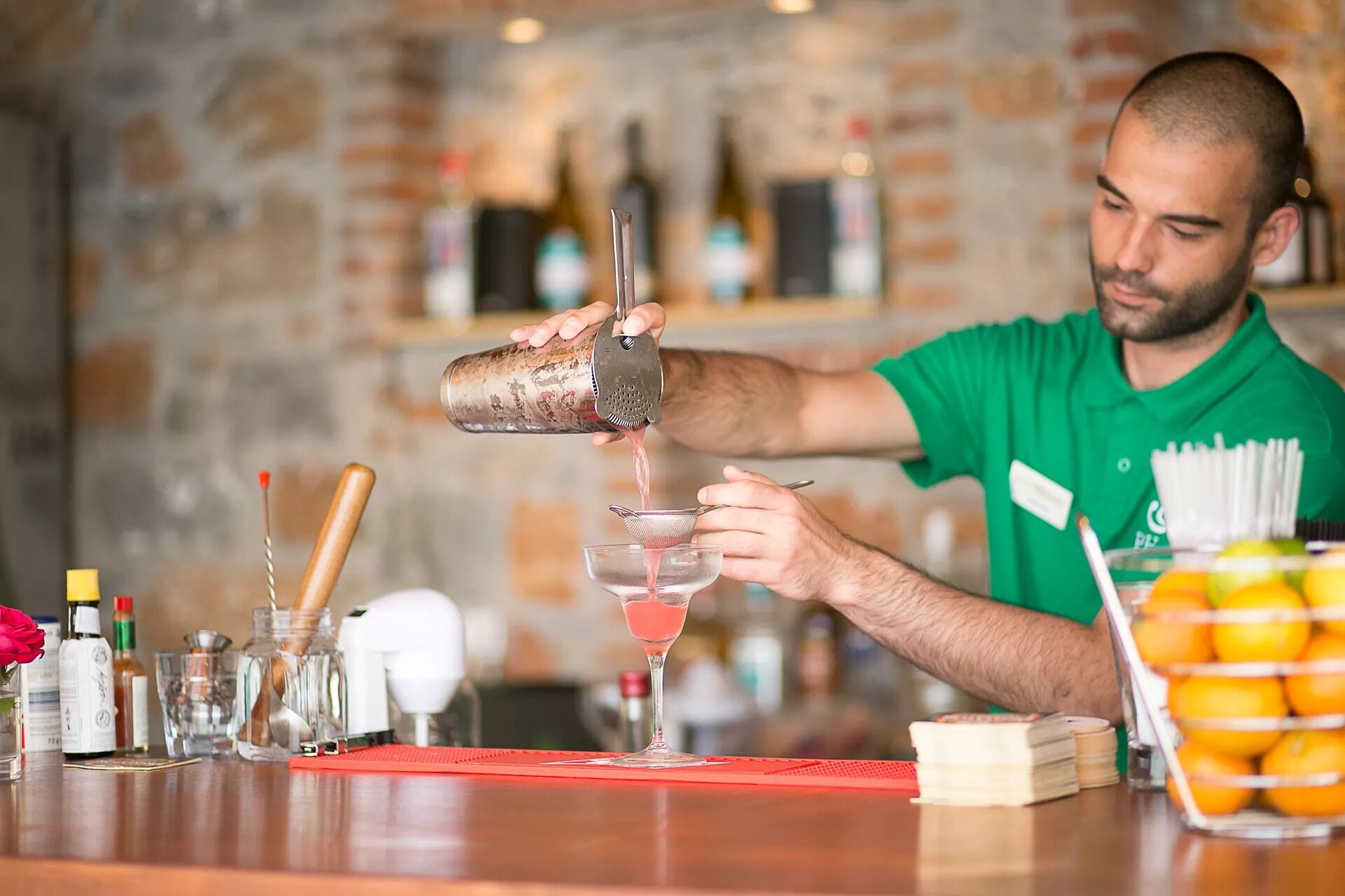 Un barman prépare un cocktail dans un bar. On aperçoit, en arrière-plan, un mur en briques, ainsi que diverses bouteilles et différents verres sur le comptoir.