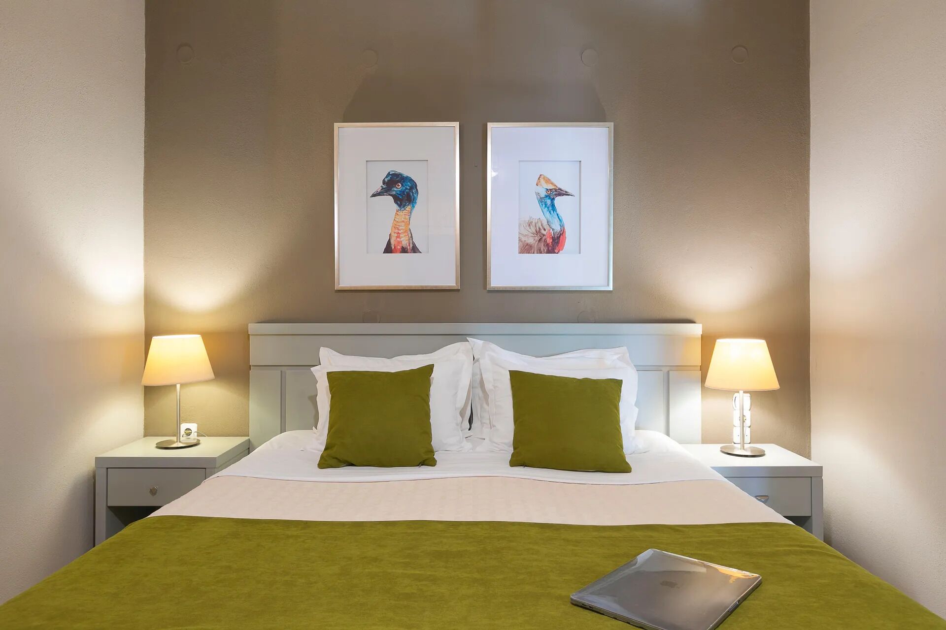 Una habitación de hotel con una cama, dos mesitas de noche, dos lámparas, una computadora portátil y dos cuadros en la pared.