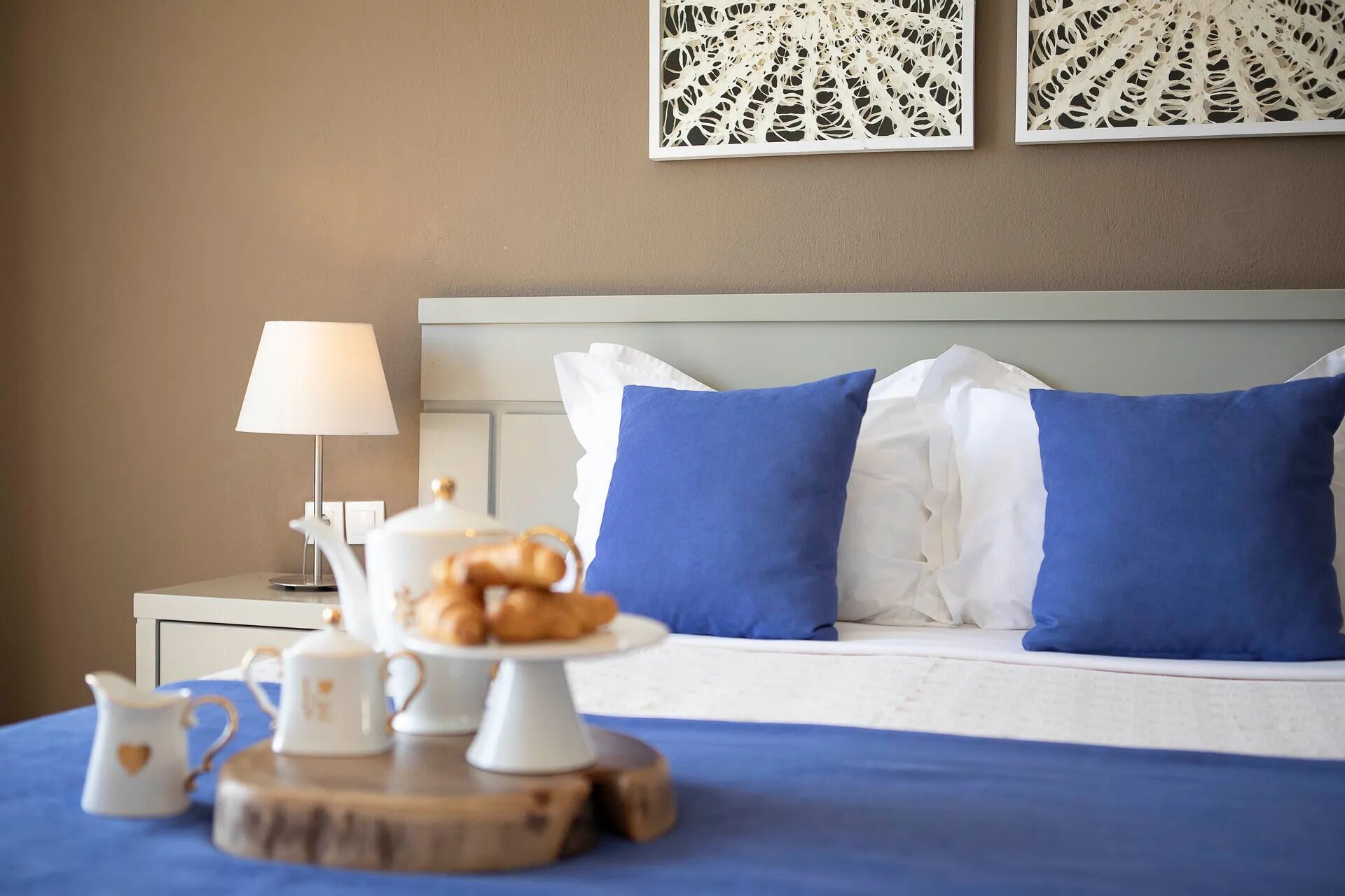 Una cama bien hecha con almohadas azules y blancas, una mesita de noche con una lámpara, y una bandeja con pasteles y teteras.