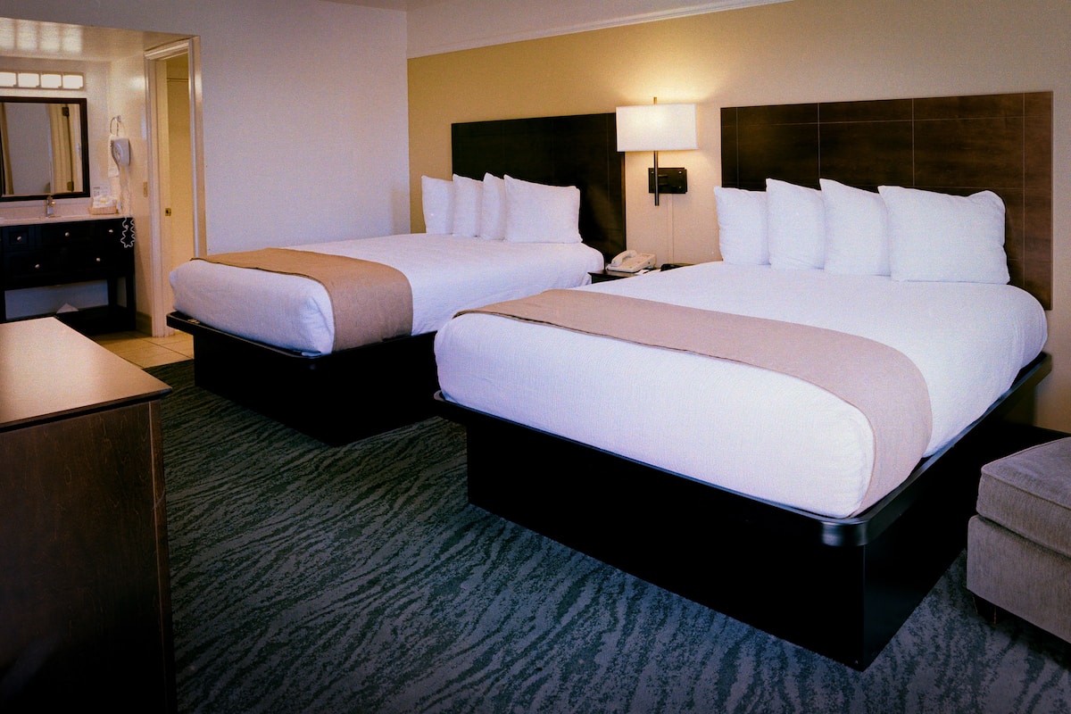 A hotel room with two beds, a desk, a mirror, and a lamp.