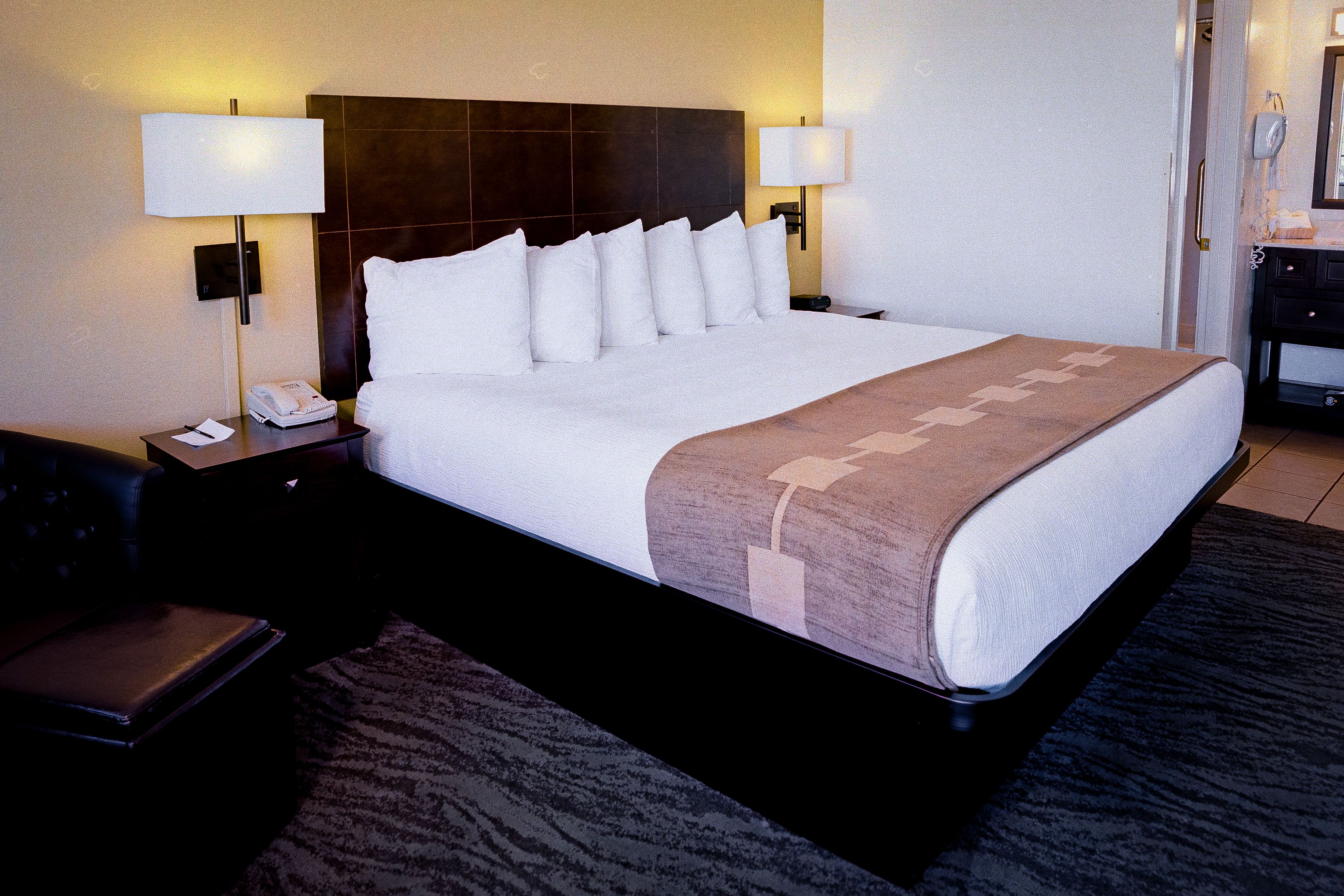 Una habitación de hotel con una cama grande, dos lámparas de noche, una mesita de noche con un teléfono y papeles, un sillón de cuero y un cabecero oscuro.