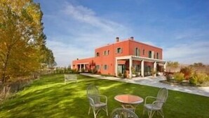 Un edificio de dos pisos con fachada roja, un patio cubierto y un césped bien cuidado con mobiliario exterior.