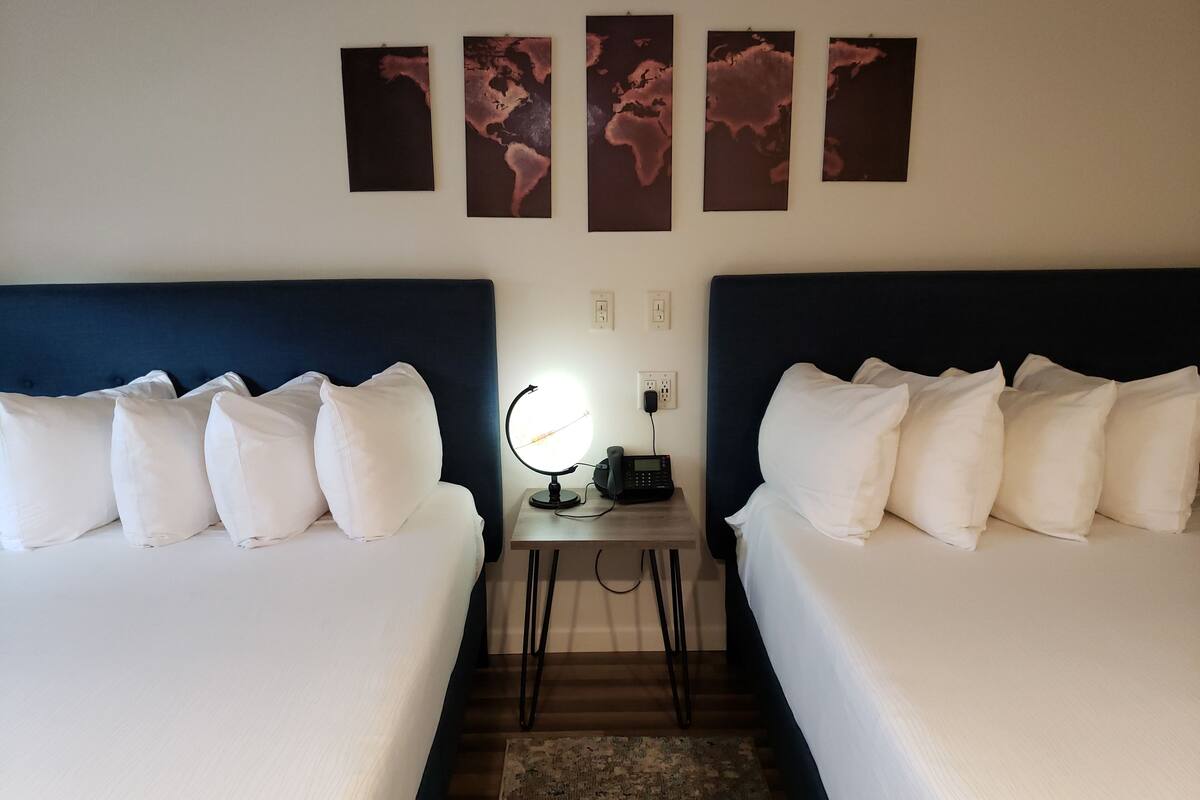 A hotel room with two beds, a nightstand with a phone and a lamp, and three world maps on the wall.