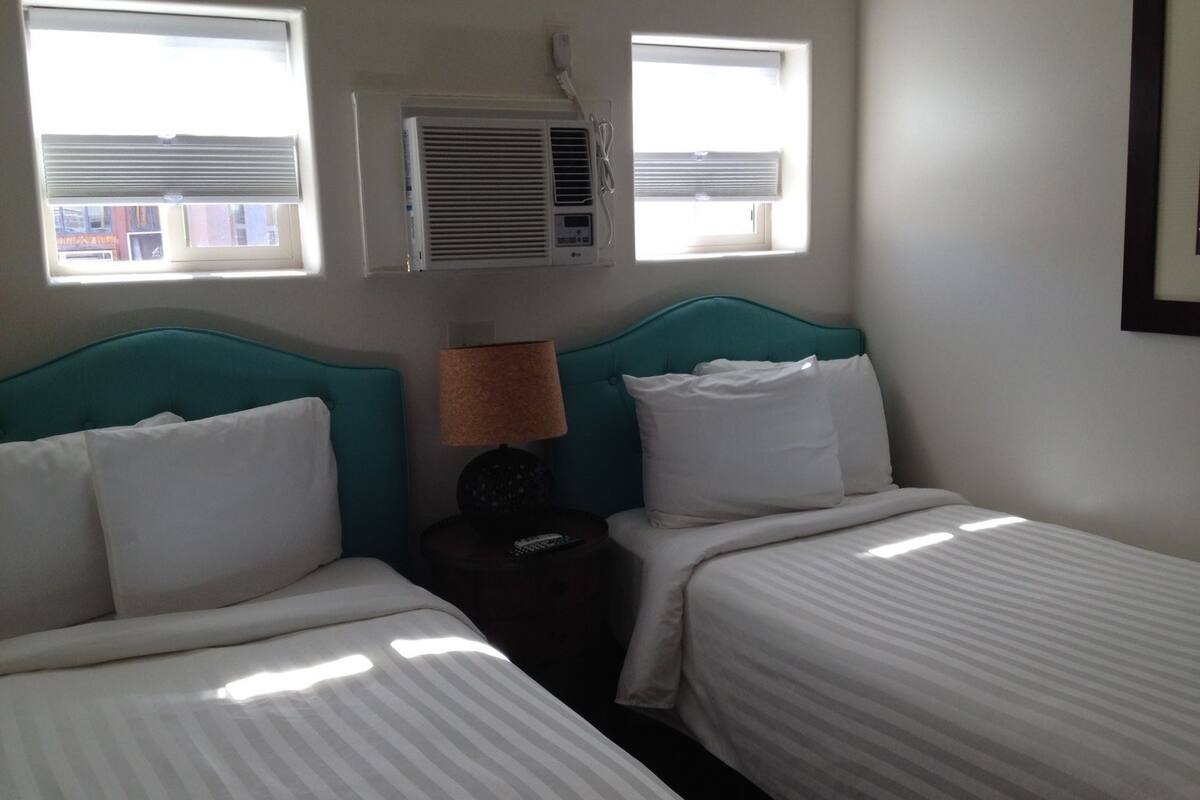 A hotel room with two beds, a window, an air conditioning unit, and a lamp on a bedside table.