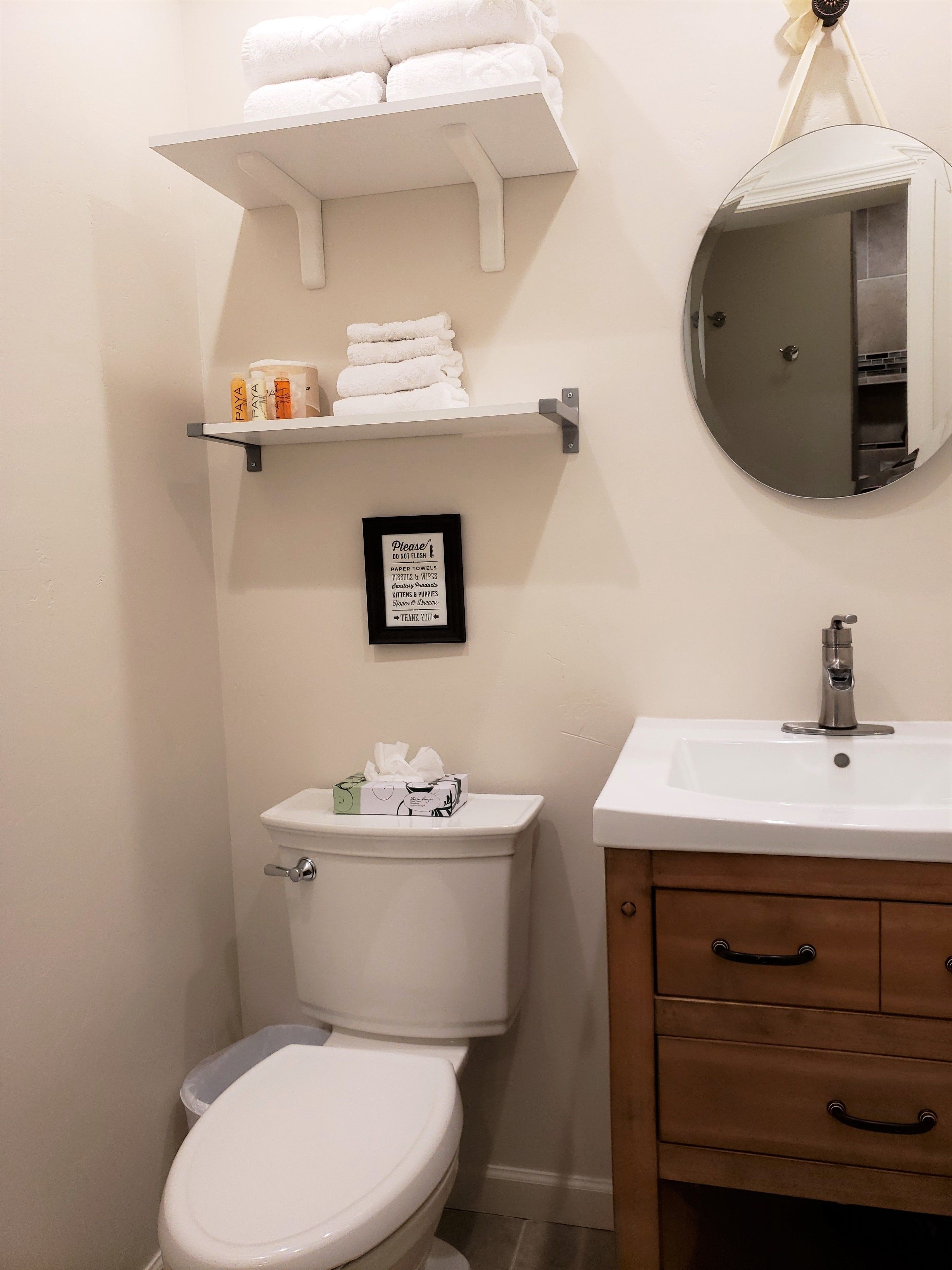A bathroom with a toilet, a sink, a vanity with drawers, a towel rack with folded towels, and a framed sign on the wall.