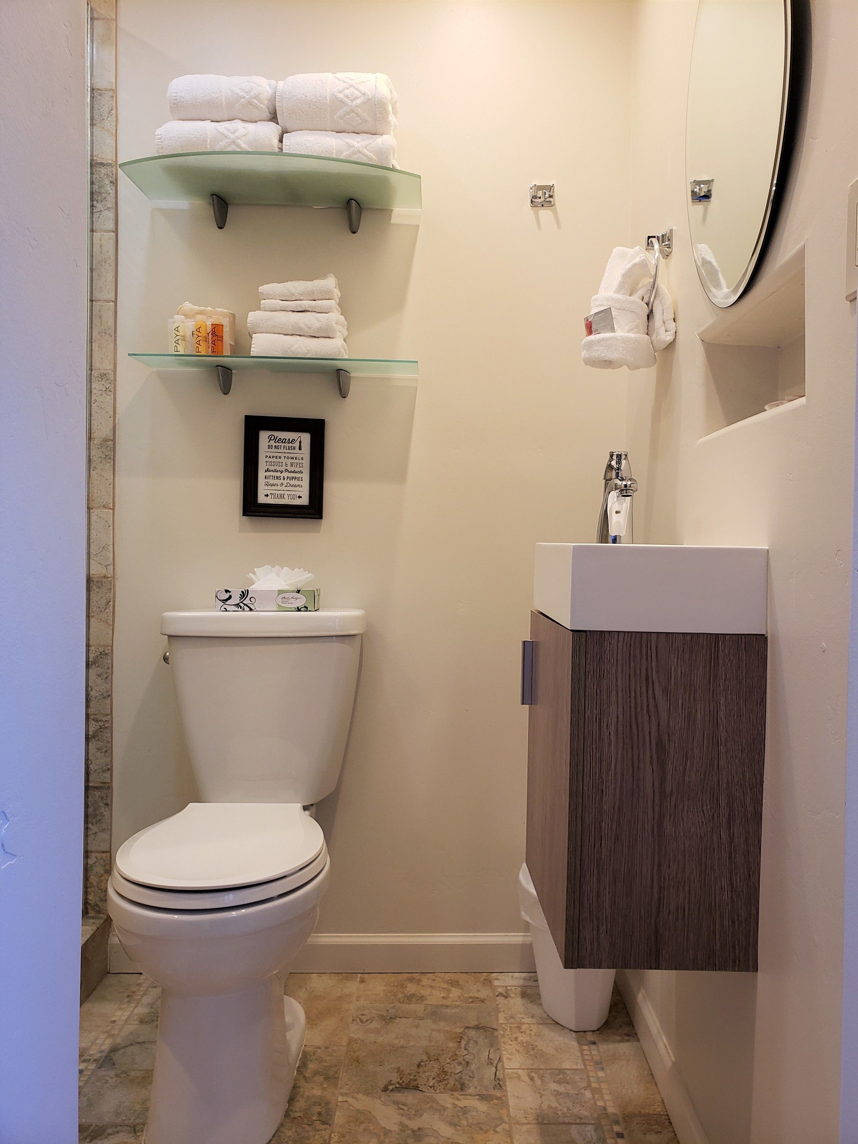 A modern bathroom with a toilet, a glass shelf with towels, a framed sign, and a wooden cabinet.
