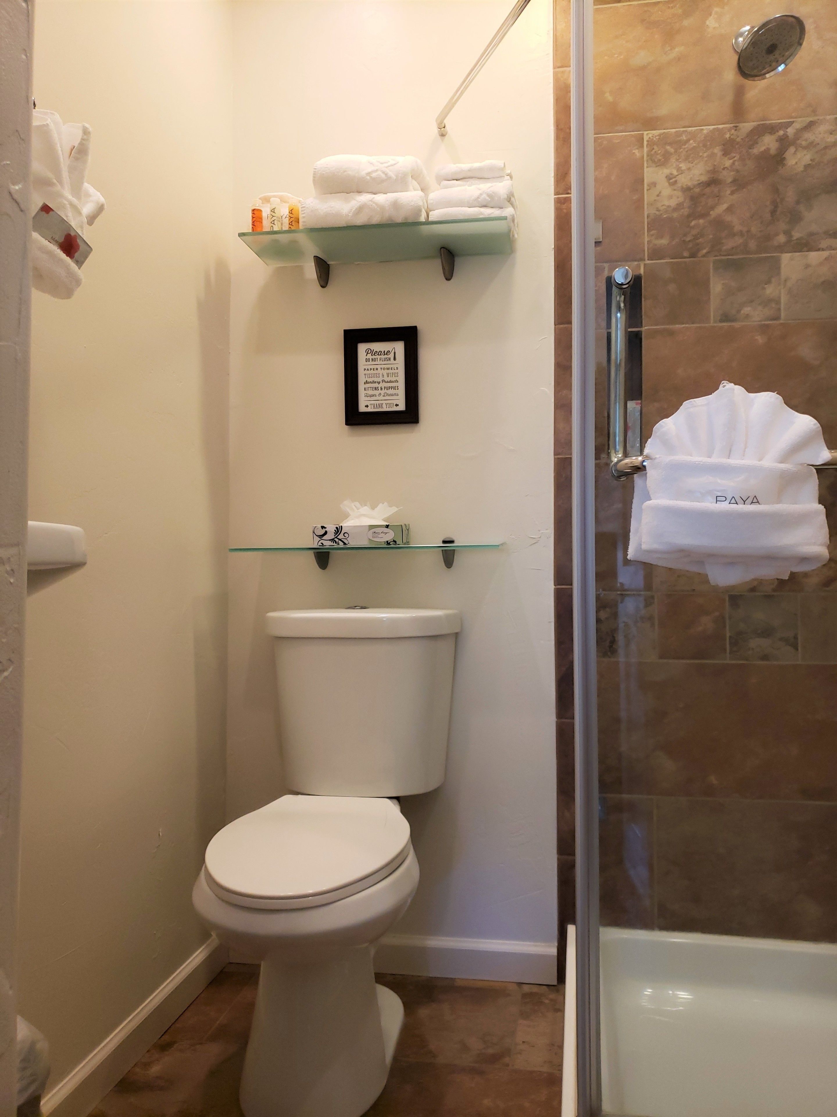 A bathroom with a toilet, a glass shelf with towels, a framed sign, and a shower area.