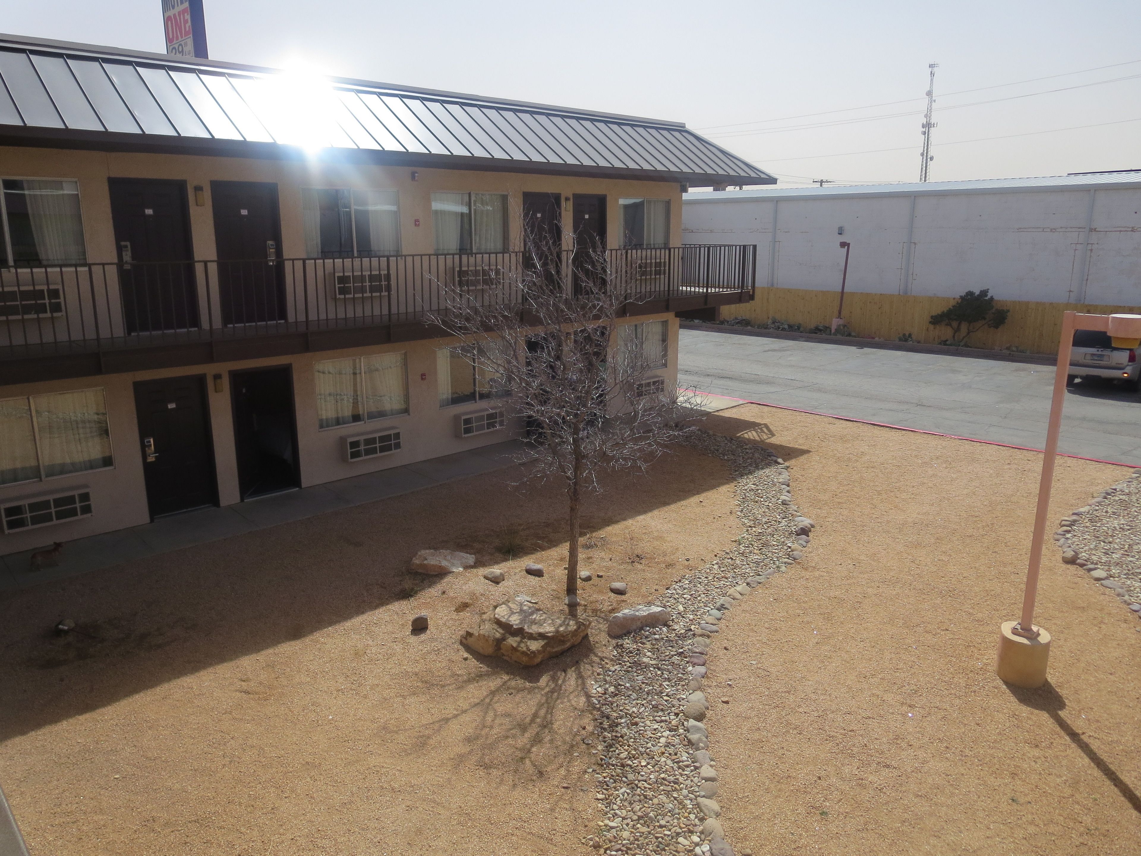 A two-story building with a metal roof and multiple windows. There is a small tree in the foreground and a paved area with a curb.