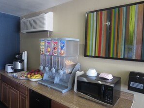 A kitchenette with a microwave, coffee maker, and a fruit bowl. There are three beverage dispensers with various drink options.