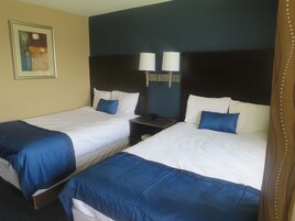 Dos camas individuales con ropa de cama blanca y azul, una mesita de noche con lámpara y un cuadro en la pared.