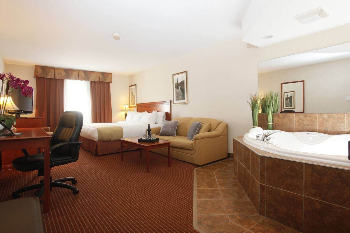 Habitación de hotel con cama, sofá, escritorio, silla, mesa de centro y baño con bañera, además de un espejo grande.