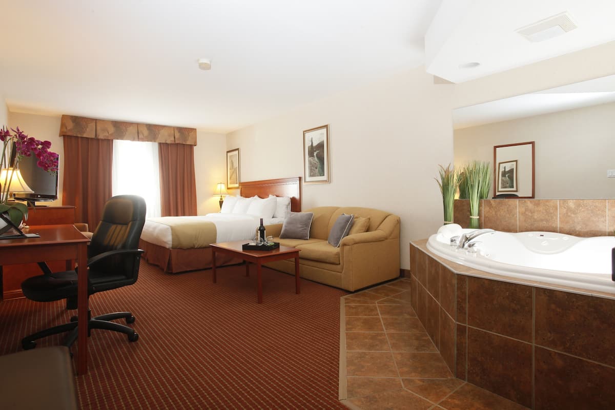 Habitación de hotel con cama, sofá, escritorio con silla, mesita, lámpara de escritorio, televisor, baño con bañera y zona de ducha.