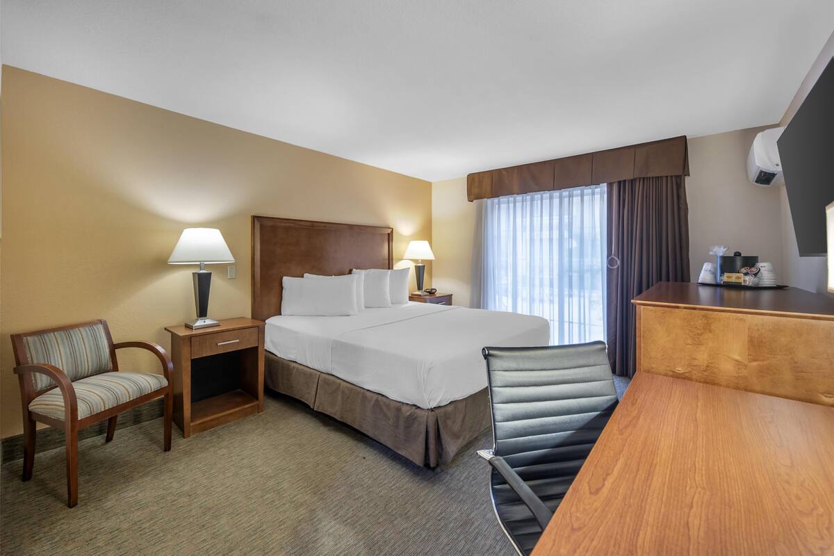 A hotel room with a bed, two bedside tables with lamps, a desk, a chair, and a large window with curtains.