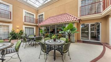 A spacious indoor patio with round tables and chairs, surrounded by potted plants and a glass ceiling.