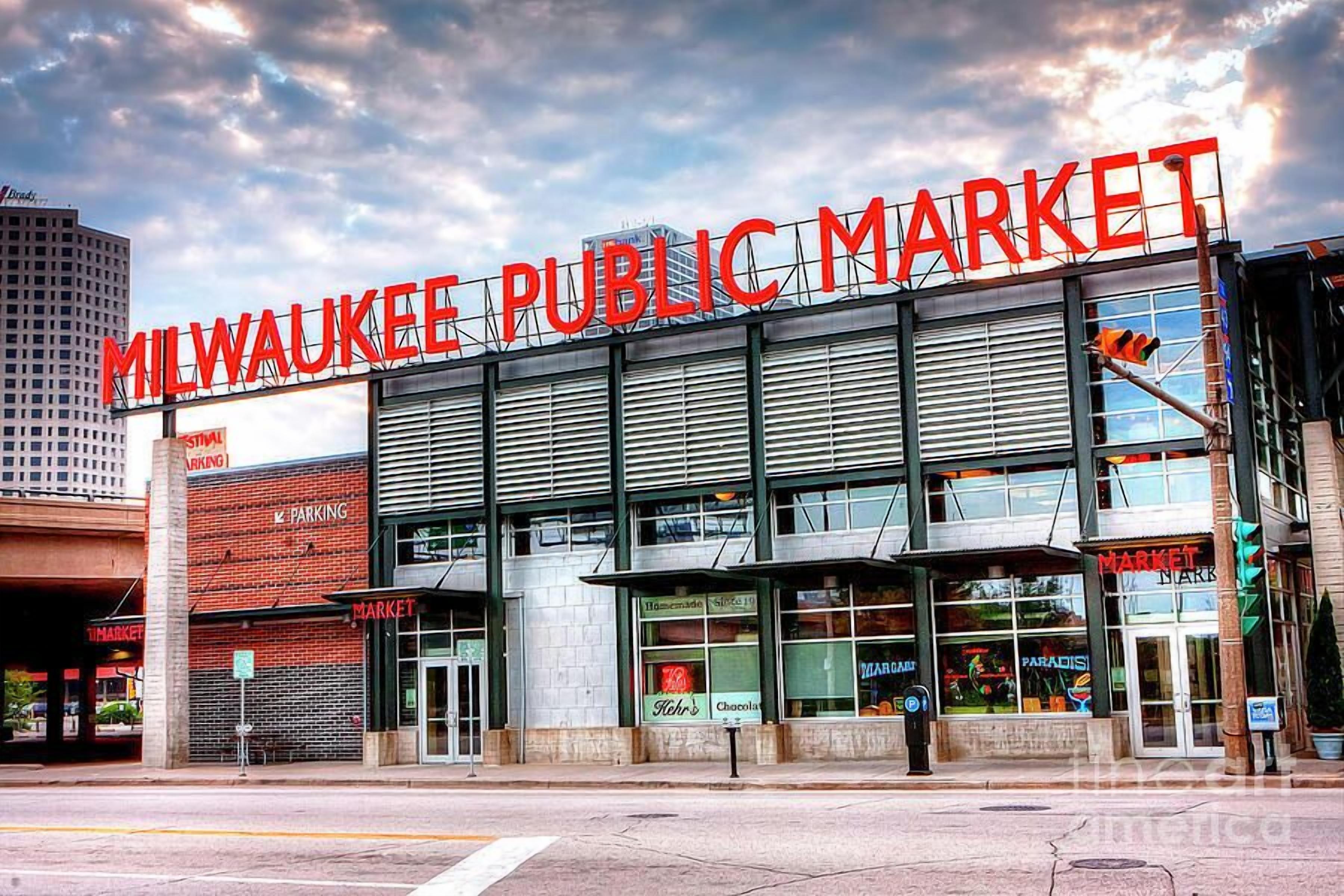 La façade du bâtiment du Milwaukee Public Market, avec ses grandes enseignes, son aire de stationnement et ses devantures de magasins.