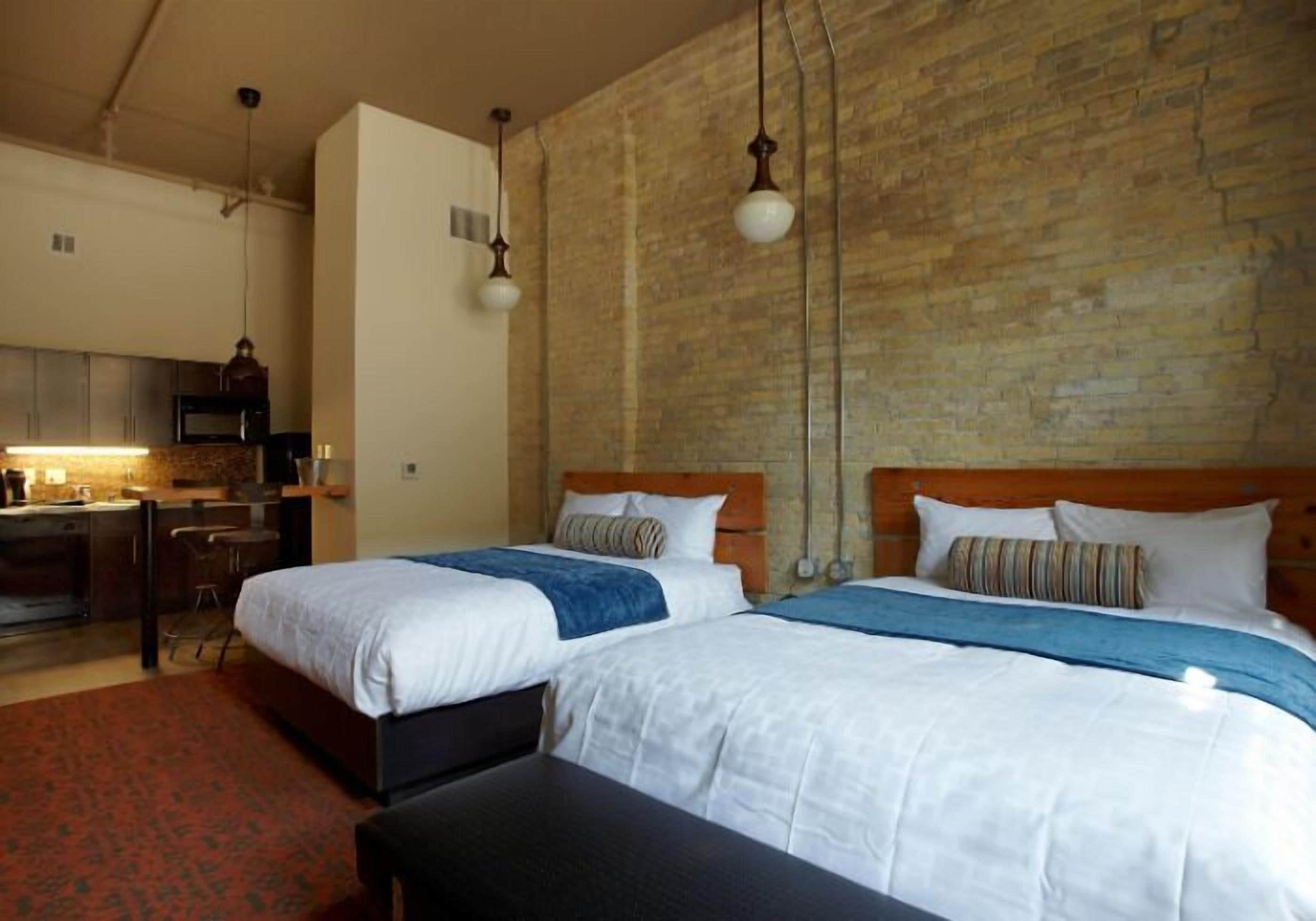 A hotel room with two beds, a kitchenette, and a brick wall.
