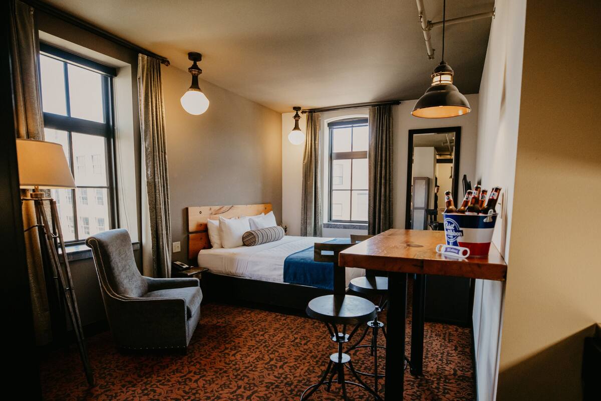 A hotel room with a bed, a desk, a chair, a lamp, and a window.